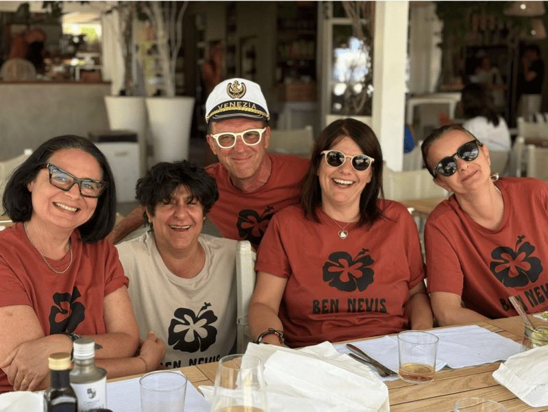 Cinque persone sedute a un tavolo di un ristorante, alcune con magliette rosse e una con maglietta bianca, sorridenti, alcune indossano occhiali da sole, e c'è una donna con cappellino. Sul tavolo ci sono bicchieri e bottiglie.