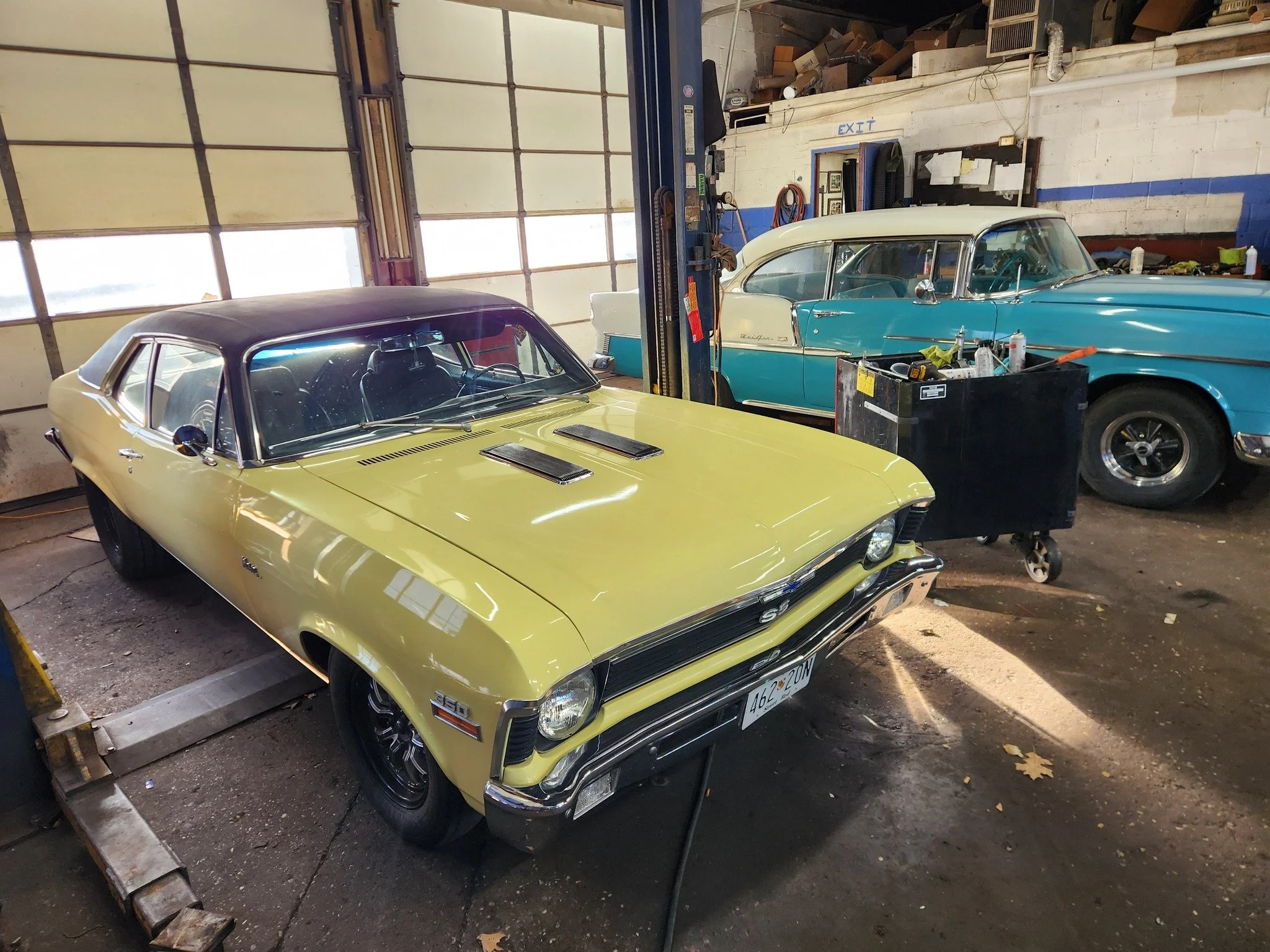 Two vintage cars inside a garage: a yellow Chevrolet Camaro SS with a black roof in the foreground, and a two-tone blue and white Cadillac in the background, with tools nearby.