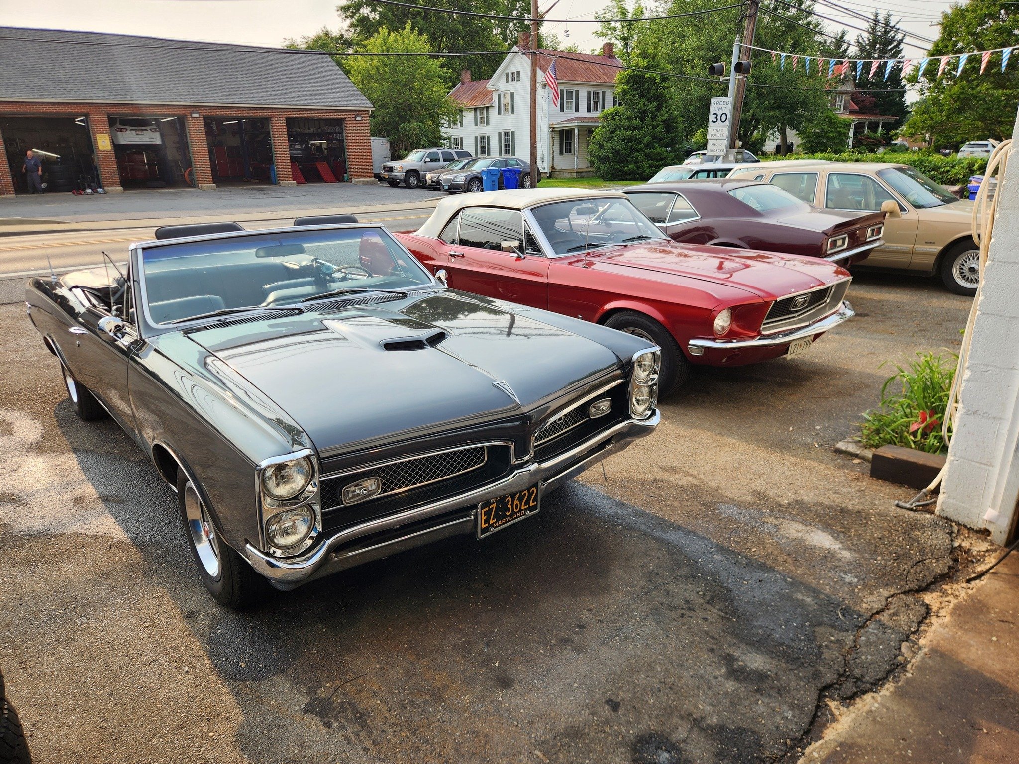 A lineup of vintage cars parked on a street, including a black convertible, a red coupe, and an older beige car, with a building and trees in the background.