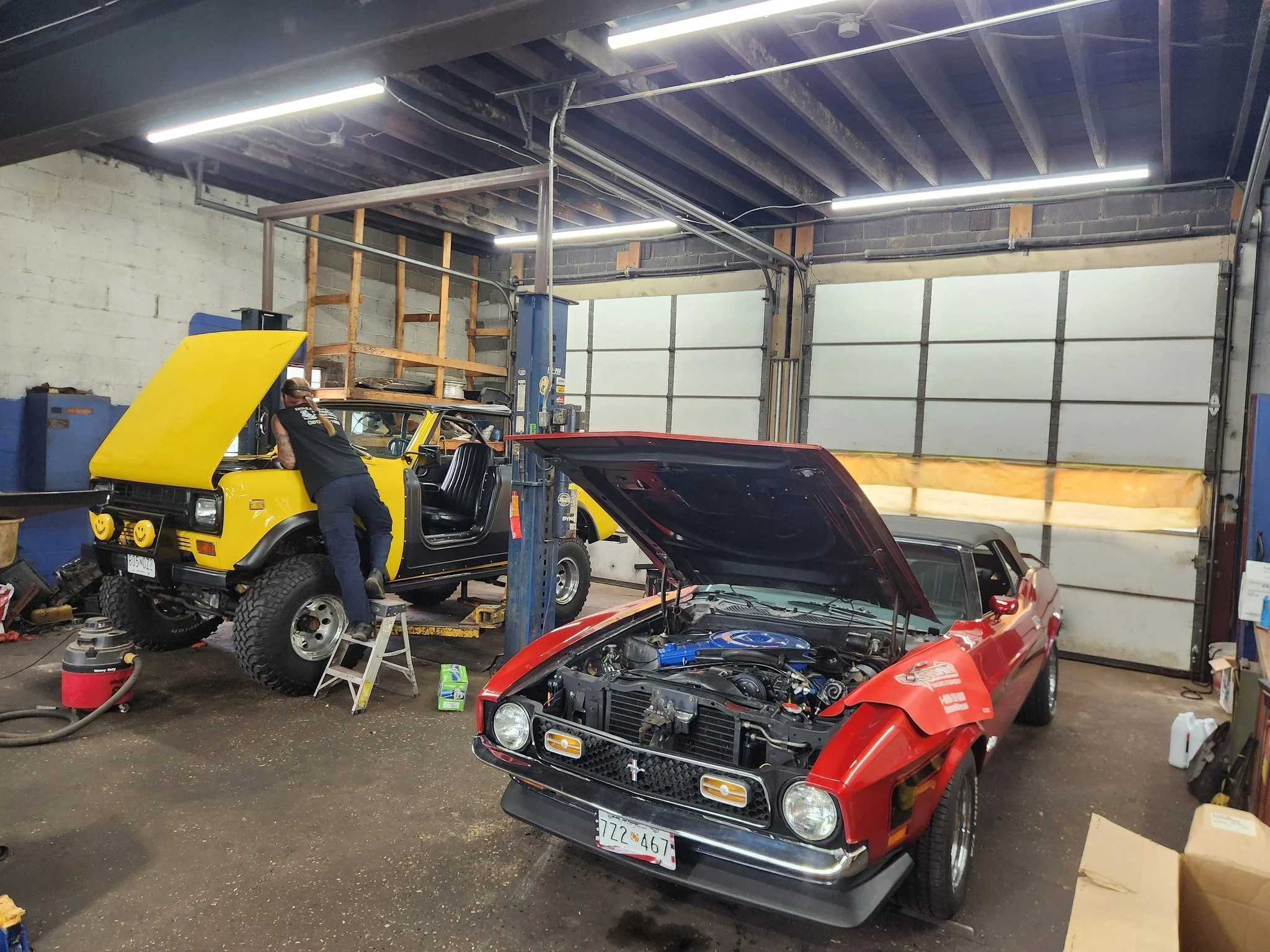 A mechanic working on a yellow off-road vehicle with its hood open in a garage, with a red vintage car with its hood open parked nearby.