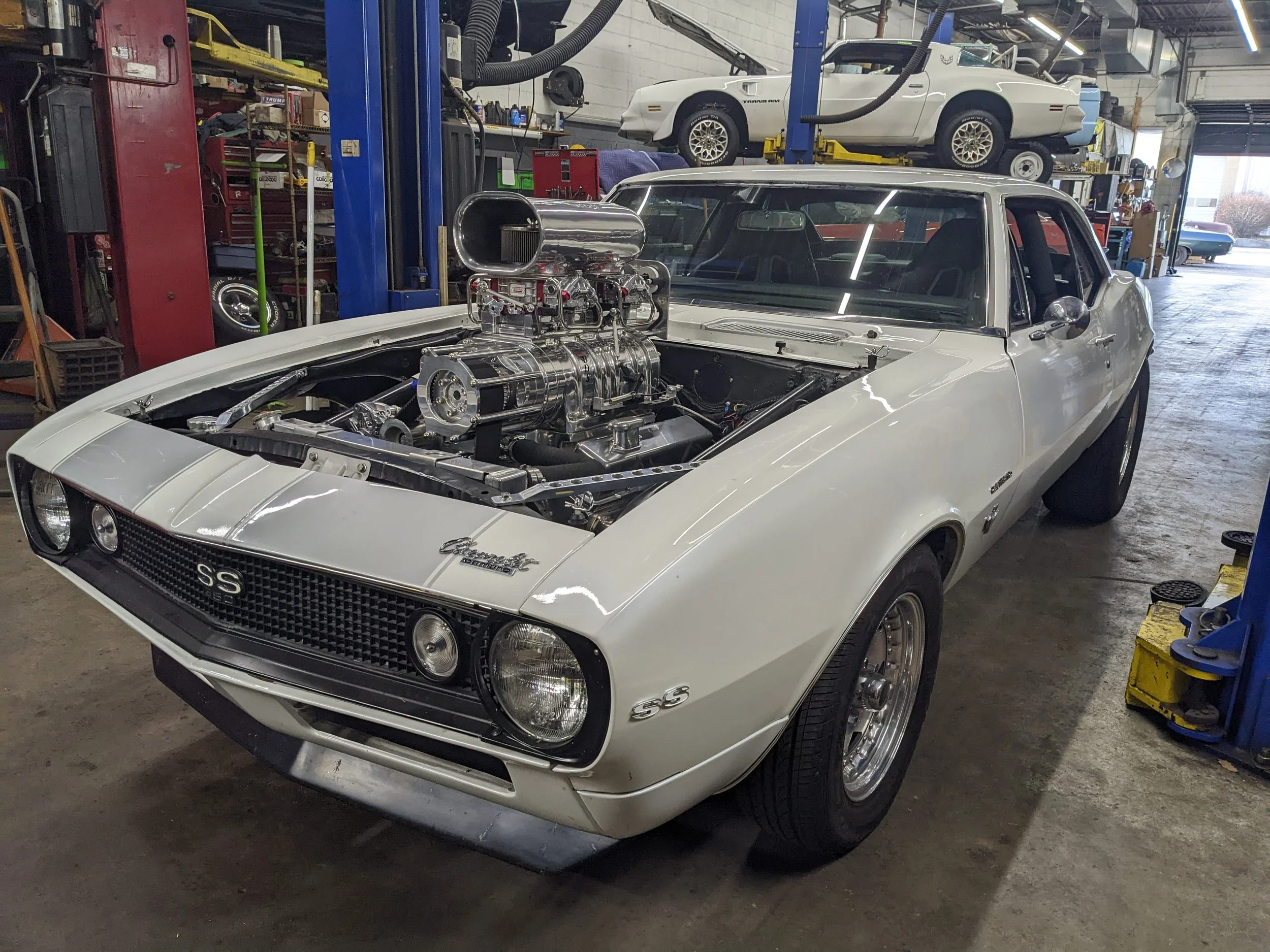 A white vintage Chevrolet Camaro SS with a supercharged engine in a garage or workshop, with various car parts and tools in the background.