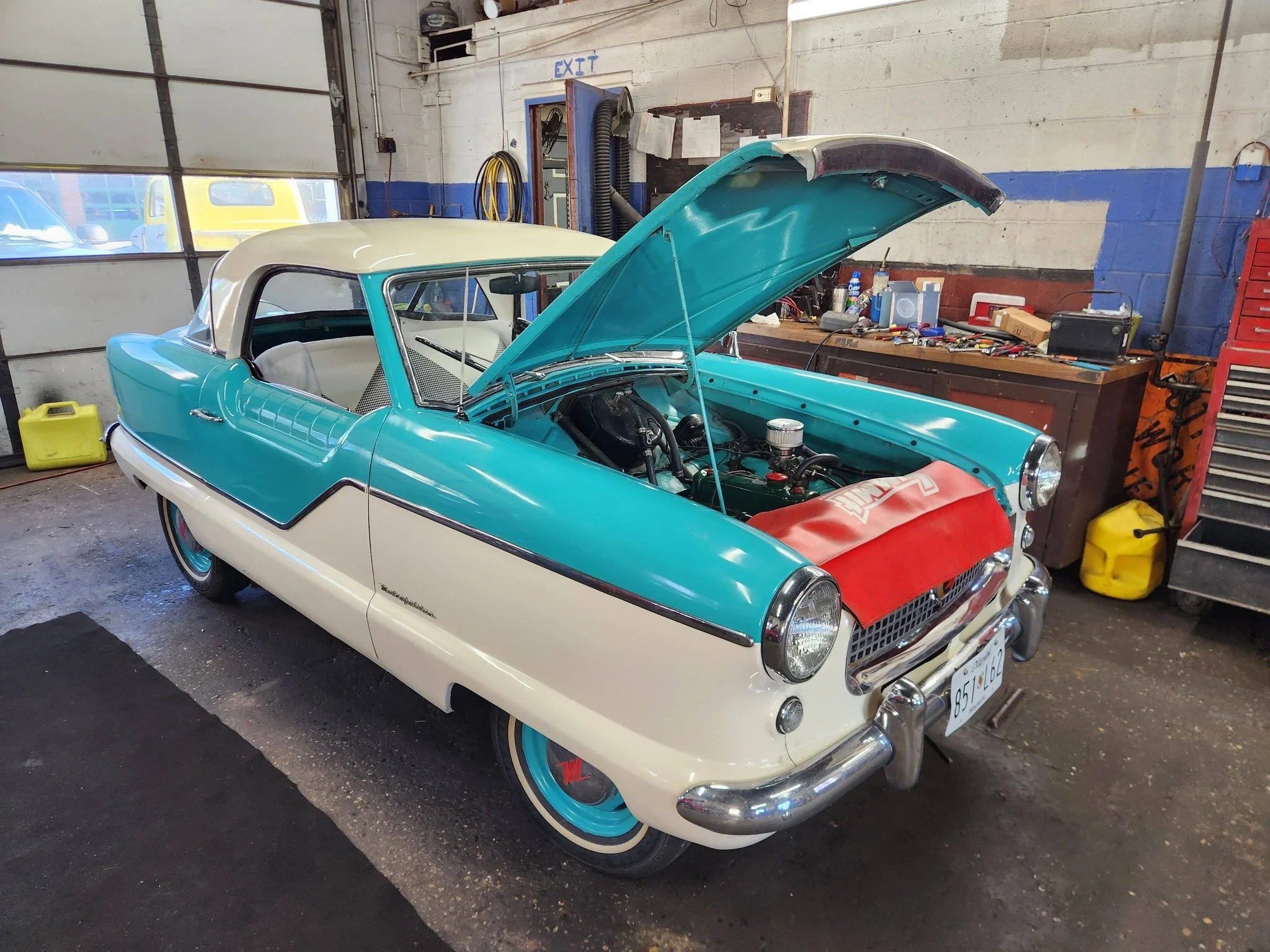 A vintage turquoise and white car with its hood open inside a garage, exposing the engine. The garage has tools on a workbench, a red tool chest, and a partially open garage door with yellow vehicles visible outside.