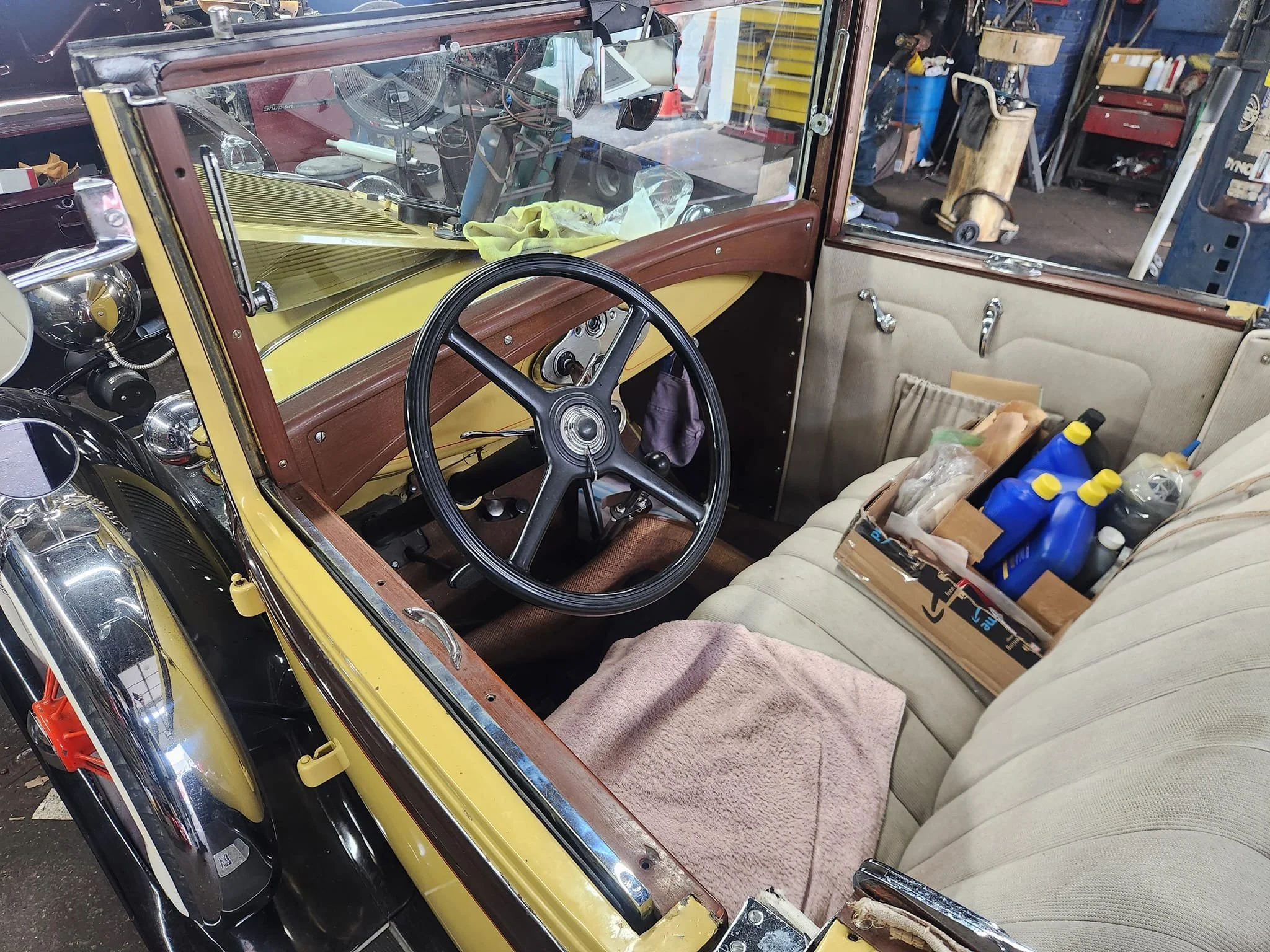 Interior of a vintage car showing the steering wheel, dashboard, front bench seat with a towel, and a cardboard box filled with cleaning supplies on the back seat, inside a garage.