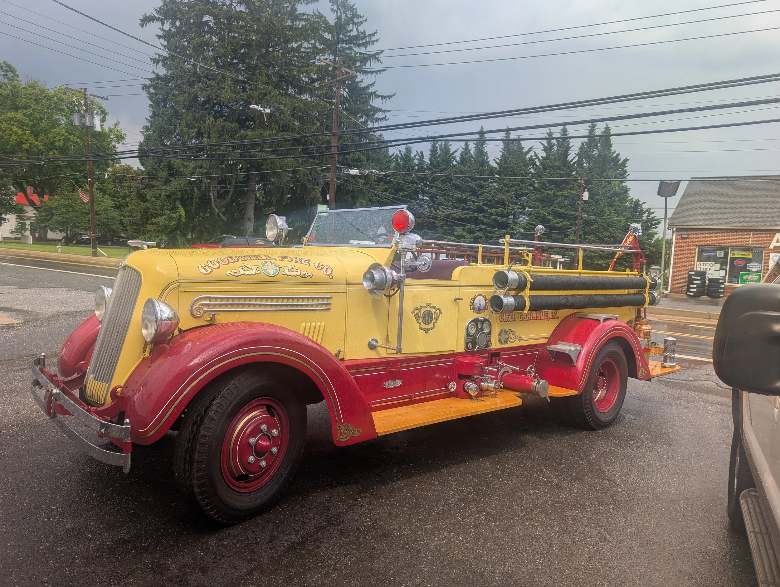 Vintage yellow and red fire engine truck parked on paved surface with trees, power lines, and buildings in the background.