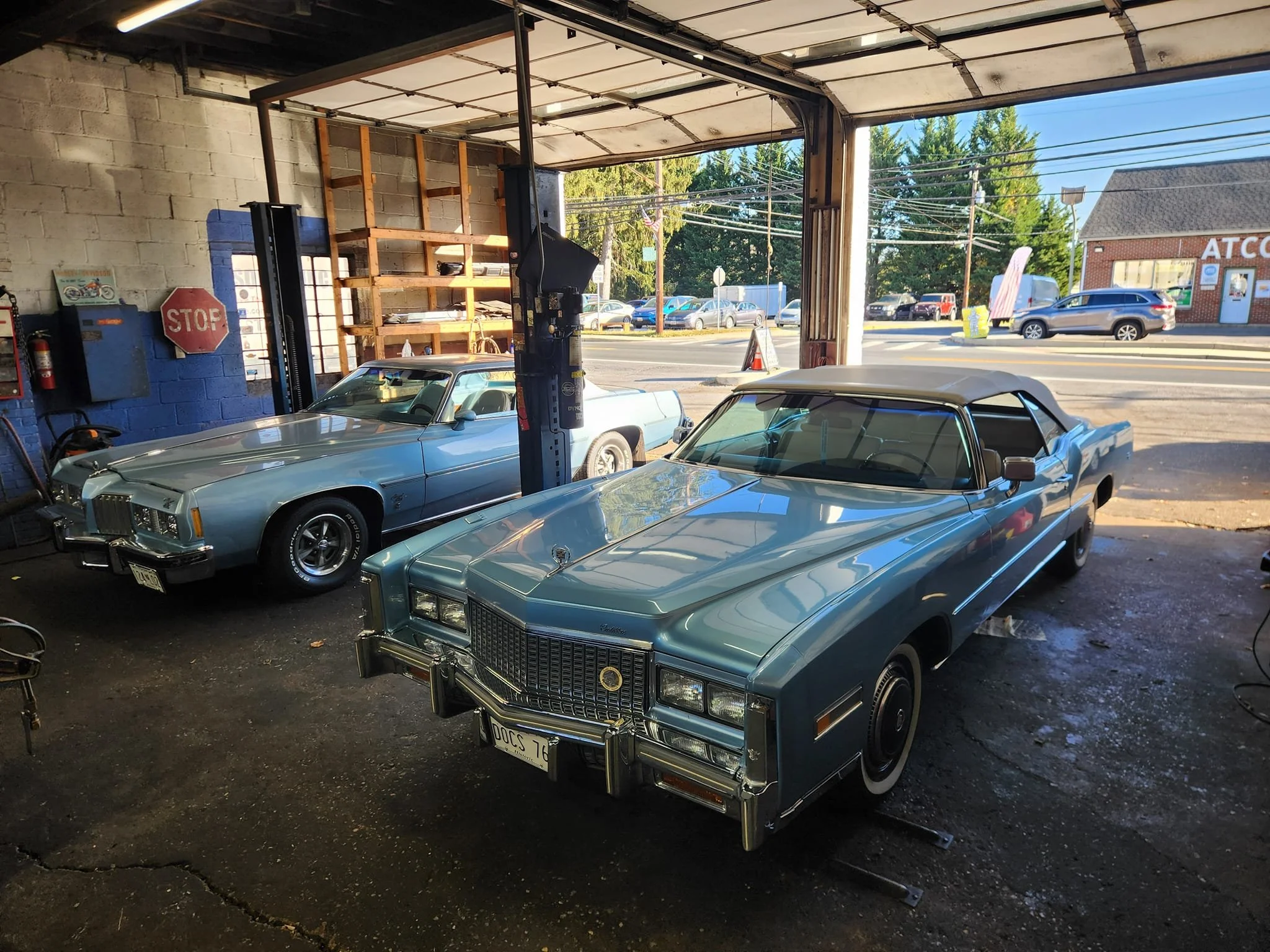 Two vintage American cars, a light blue 1970s Cadillac and a silver 1970s Pontiac, parked in a garage with an open front, allowing a view of the street outside.