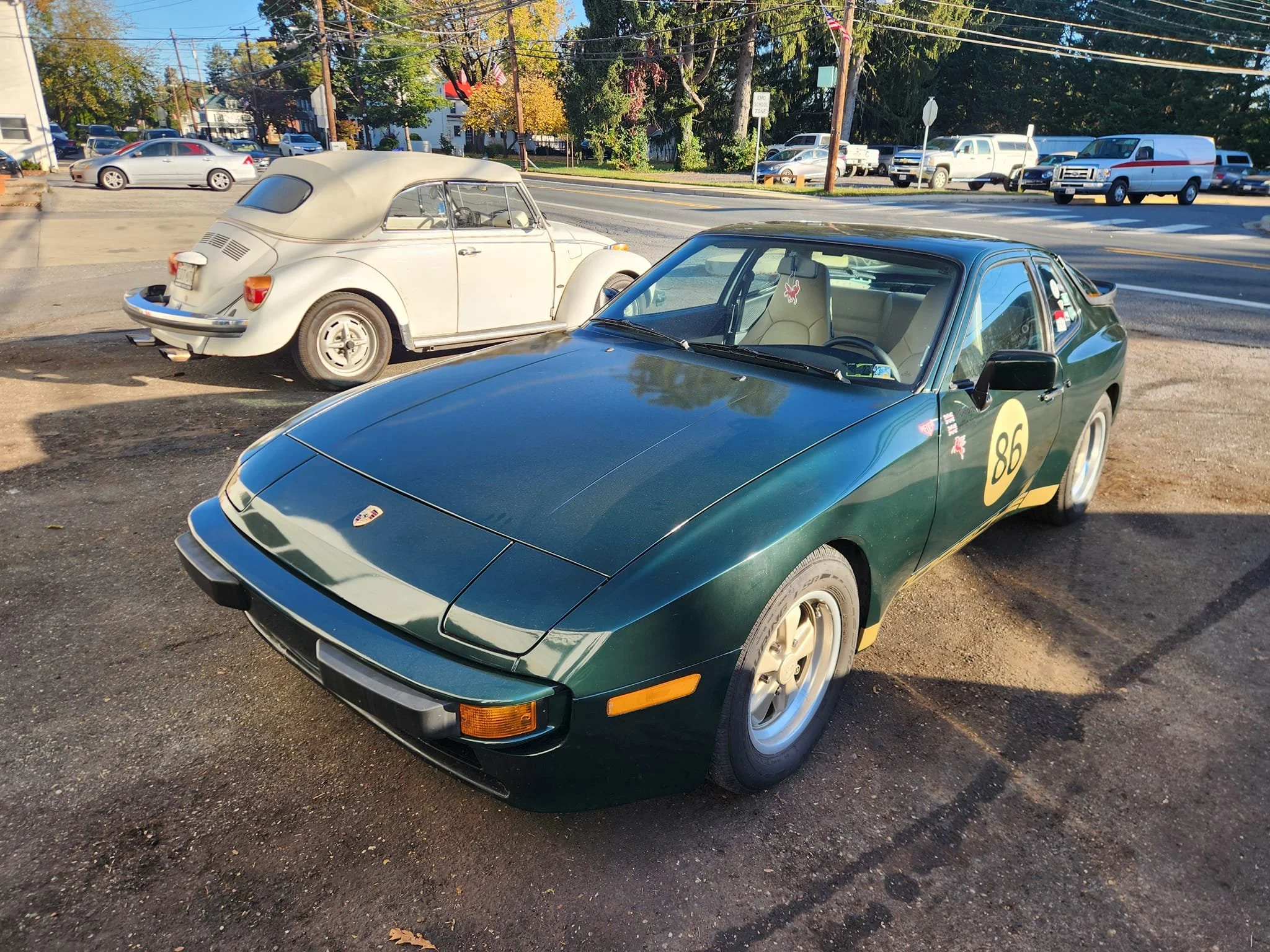A green vintage Porsche sports car with a yellow circle and the number 86 on the door, parked on a dirt lot next to a white classic Volkswagen Beetle, with a busy street and cars in the background.