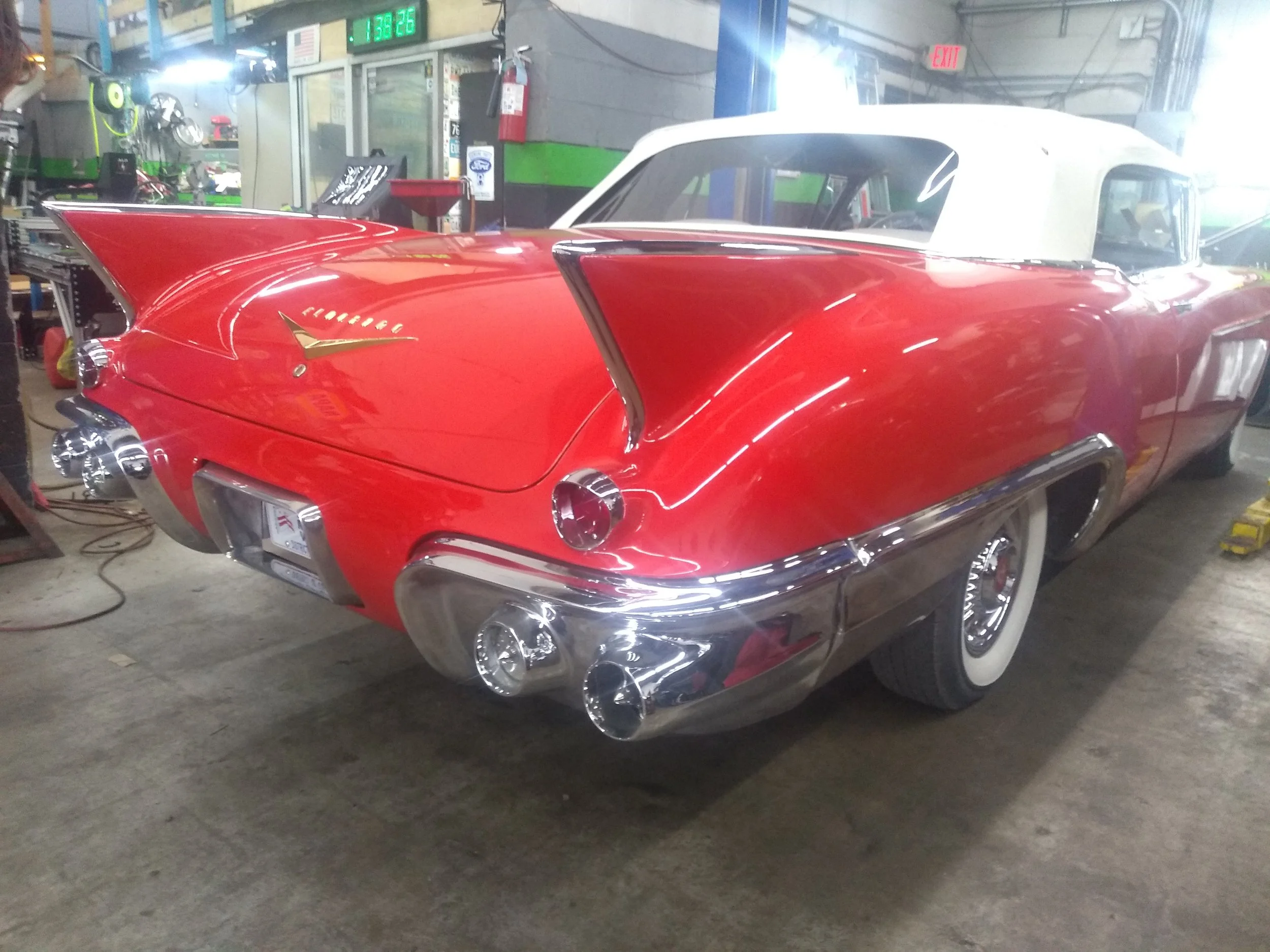 A vintage red Cadillac convertible with a white roof, displaying distinctive tail fins and chrome details, parked indoors in a mechanic's garage.