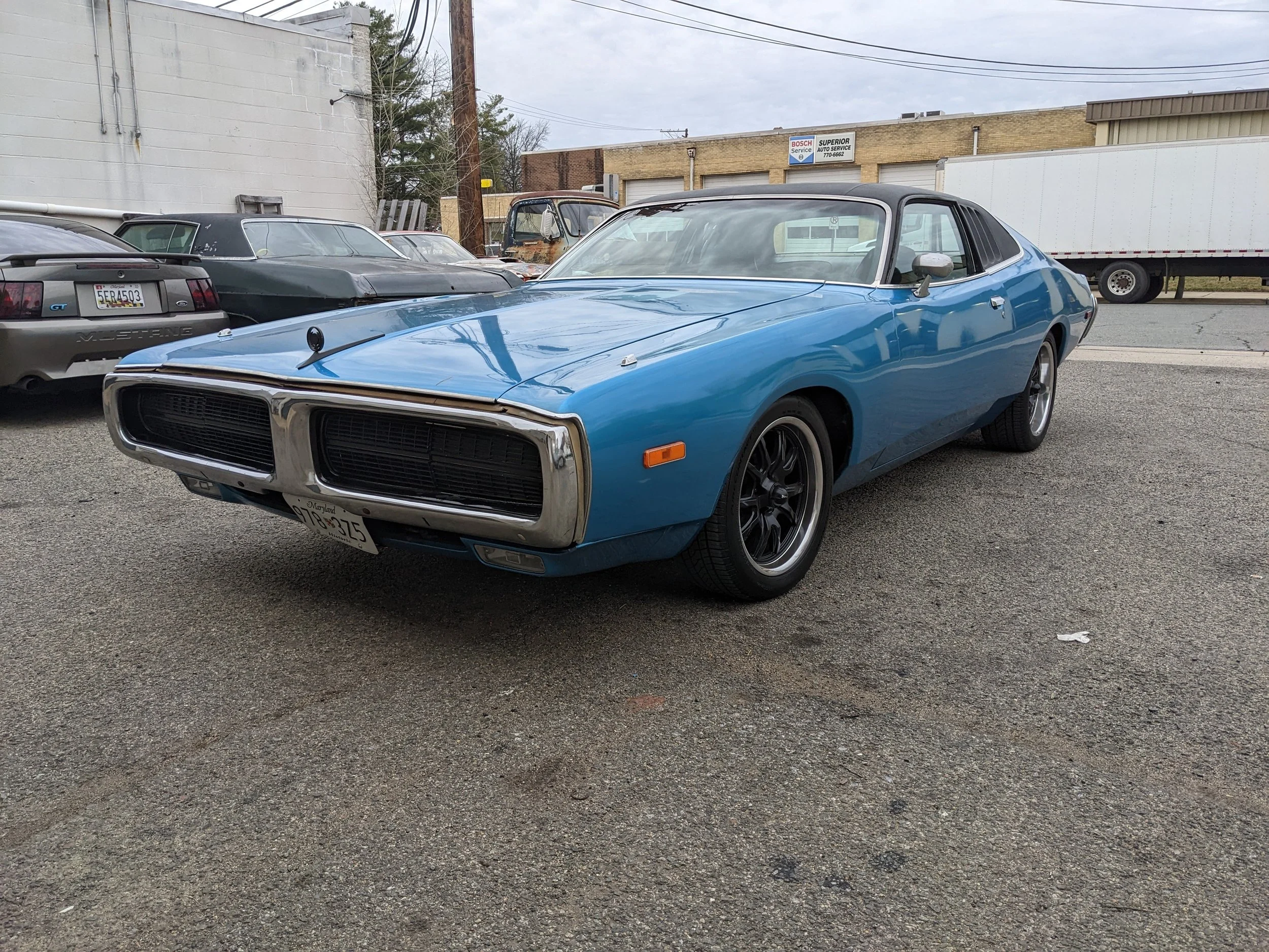 Blue vintage Dodge Charger muscle car parked in an outdoor lot with other classic and modern vehicles, industrial buildings, and trucks in the background.