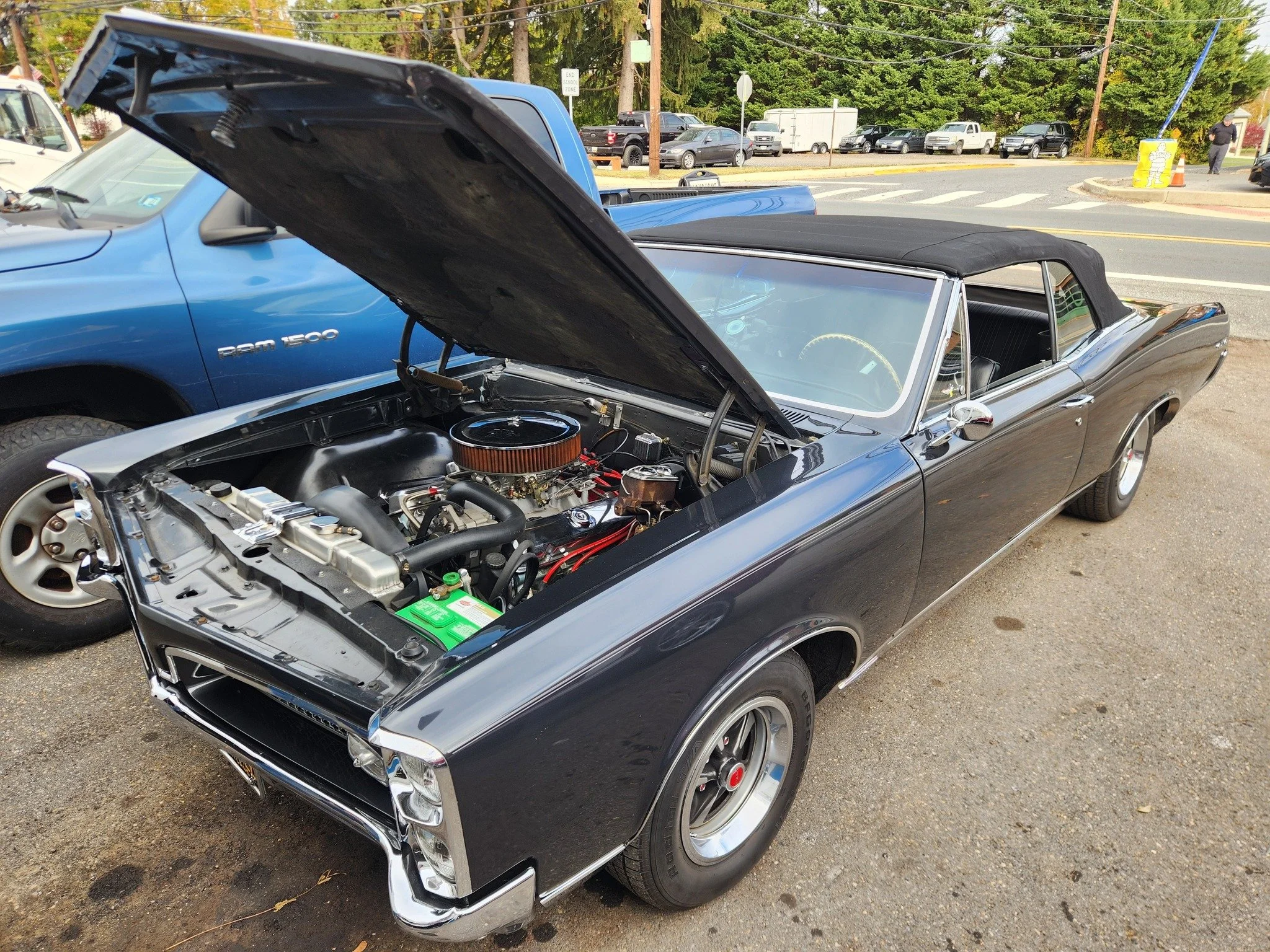 Black classic convertible car with open hood showing engine, parked on a street with other vehicles and trees in the background.