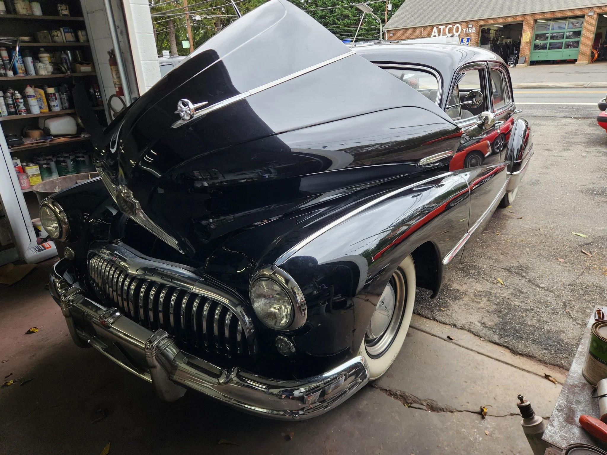 Vintage black and red car with a streamlined design parked inside a garage with tools and supplies on shelves, with a view of a street and a building labeled ATCO TIRE outside.