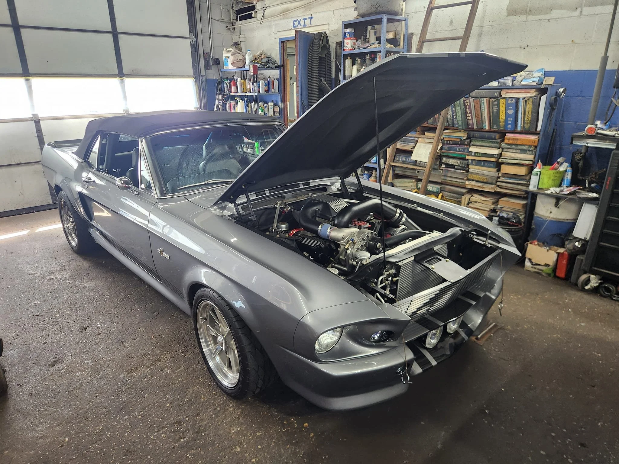 A gray classic Ford Mustang in a garage with its hood open, revealing a modified engine. The garage has shelves filled with books and tools.