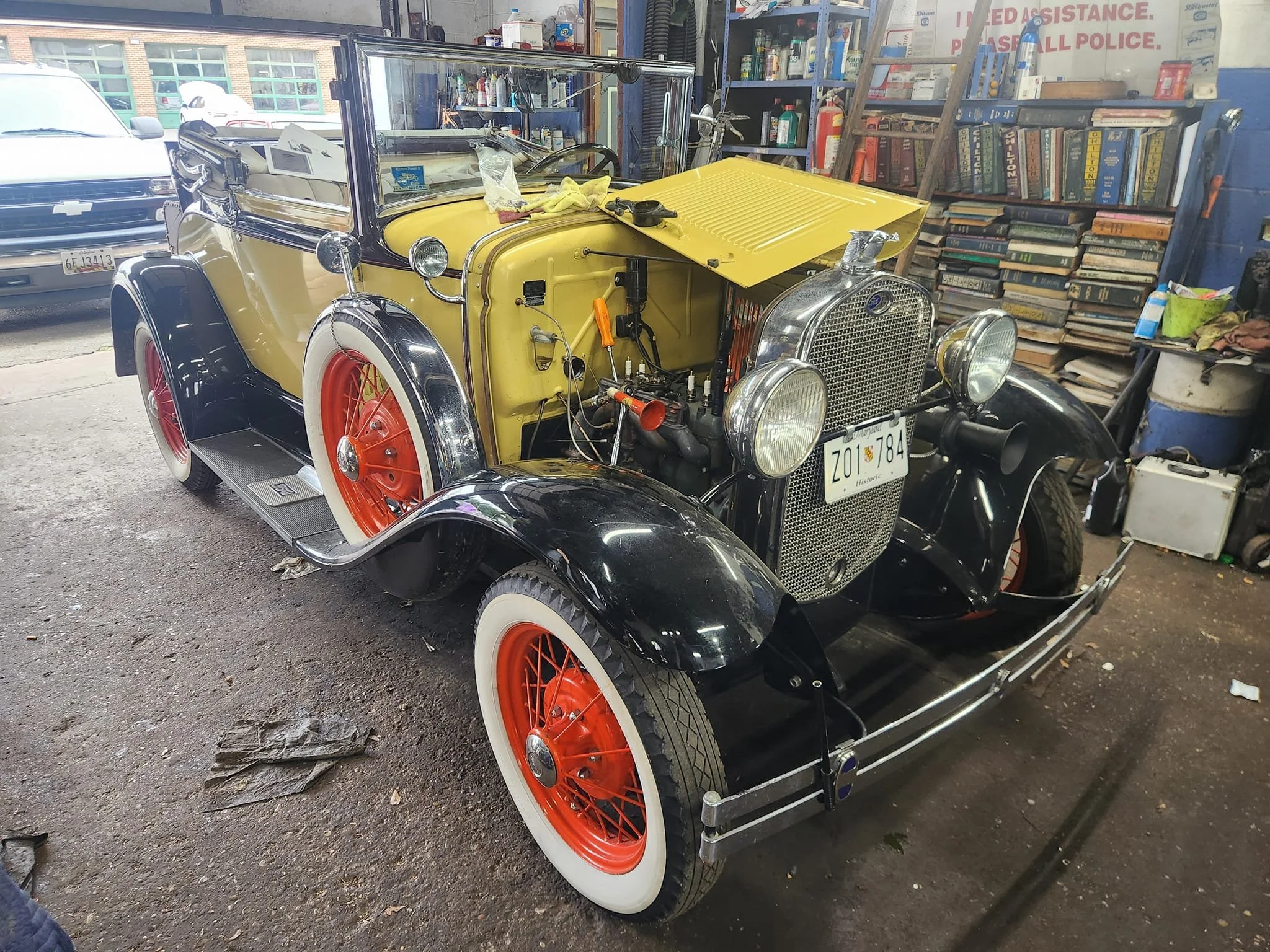 A vintage yellow-and-black classic car inside a garage, with an open hood, red wheels with white tires, and various tools and shelves in the background.