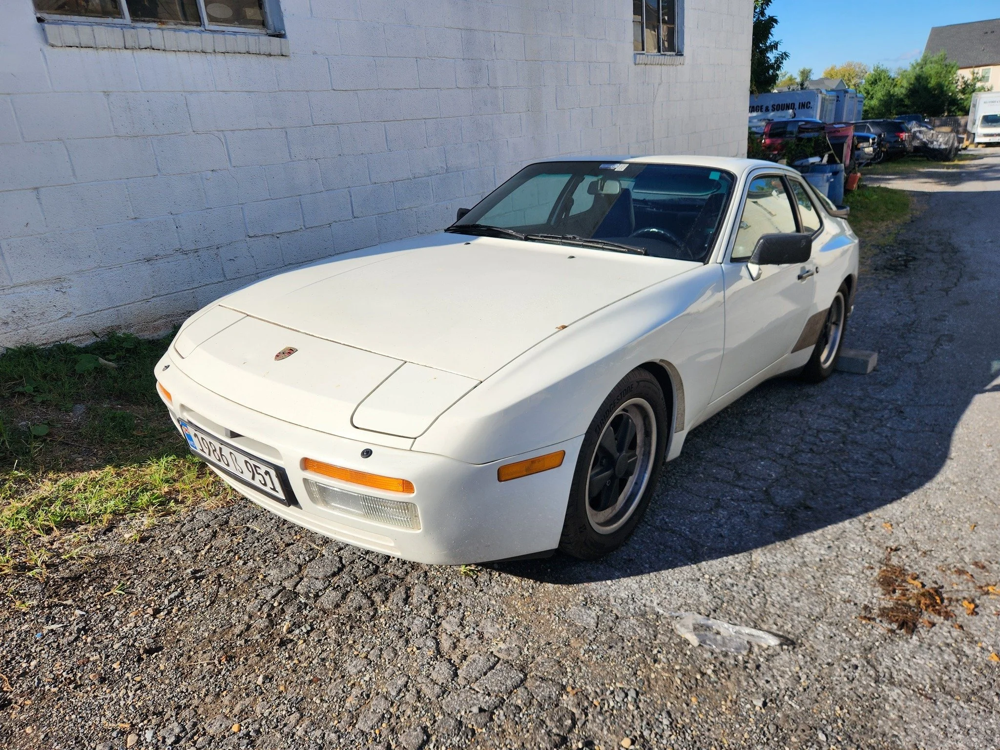 A white vintage Porsche 944 sports car parked on a gravel lot next to a white brick building, with other vehicles and trees in the background.