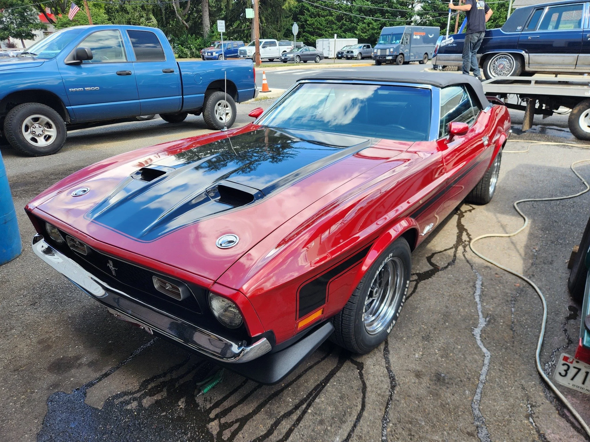 Red vintage muscle car with black racing stripes parked at a car wash, with a blue pickup truck and other vehicles in the background.