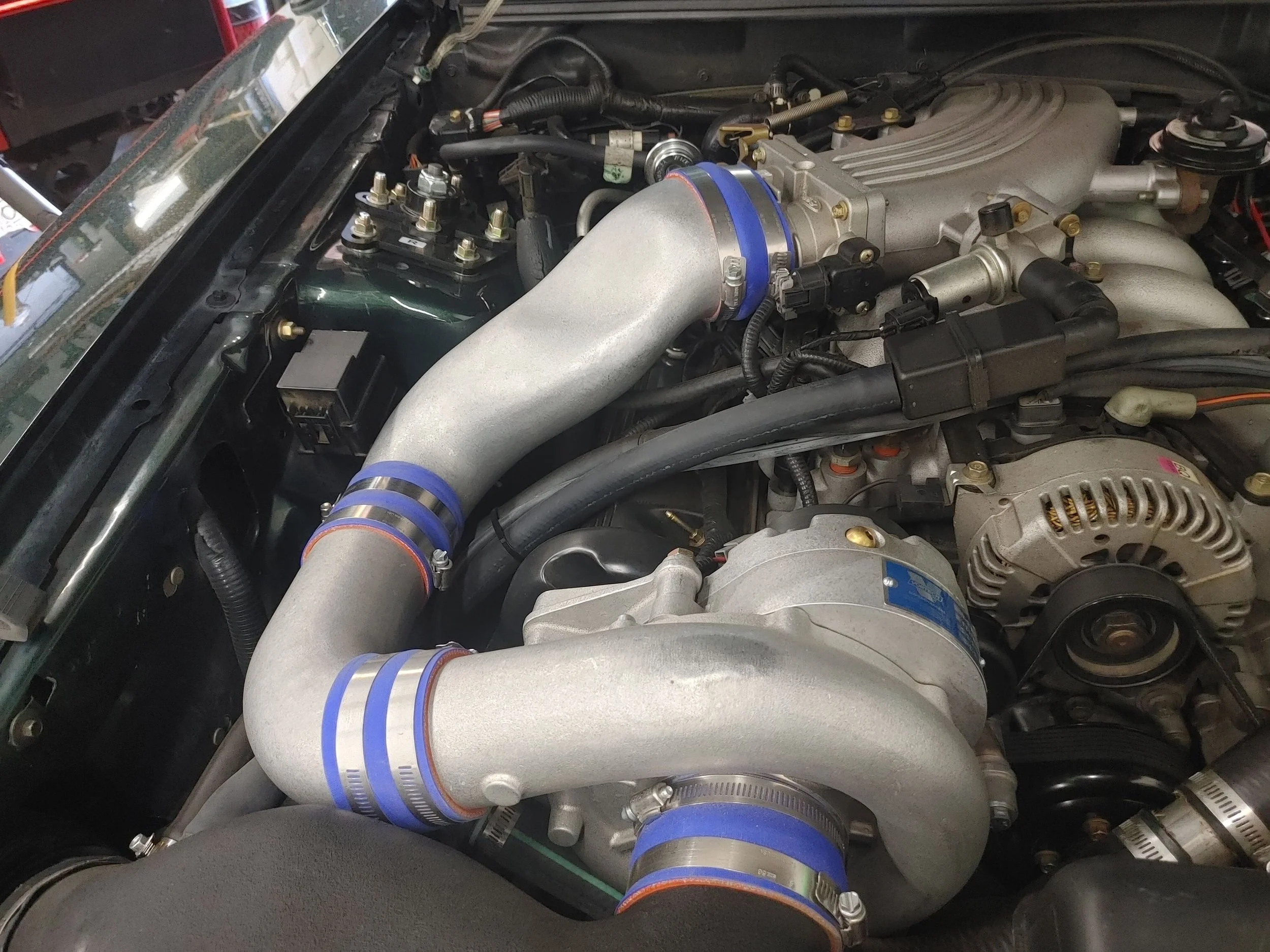 Close-up of a car engine with large silver turbocharger piping, blue silicone couplers, and various engine components including an alternator, wiring, and metal brackets.