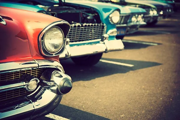 Close-up of vintage American cars parked in a parking lot, focusing on the front of a red car with chrome details and other classic cars in the background.