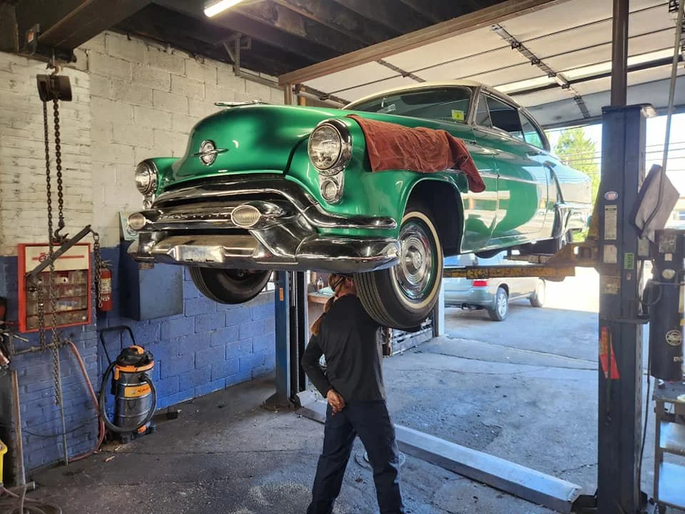 A vintage green car elevated on a hydraulic lift inside an auto repair garage, with a mechanic standing below.