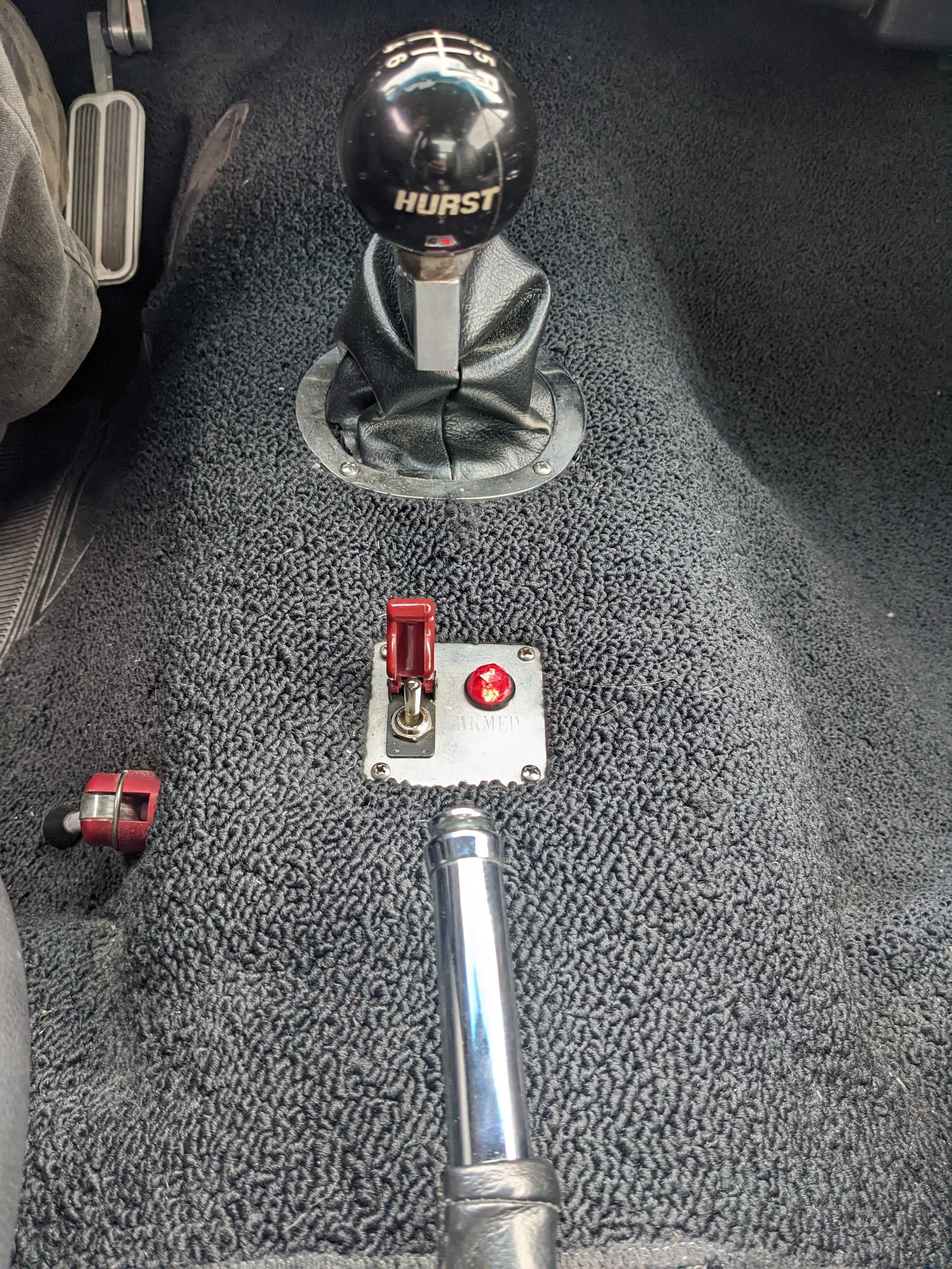 Close-up of a car's interior focusing on a Hurst gear shift lever and a toggle switch with a red indicator light near the center console, with a textured black carpet.