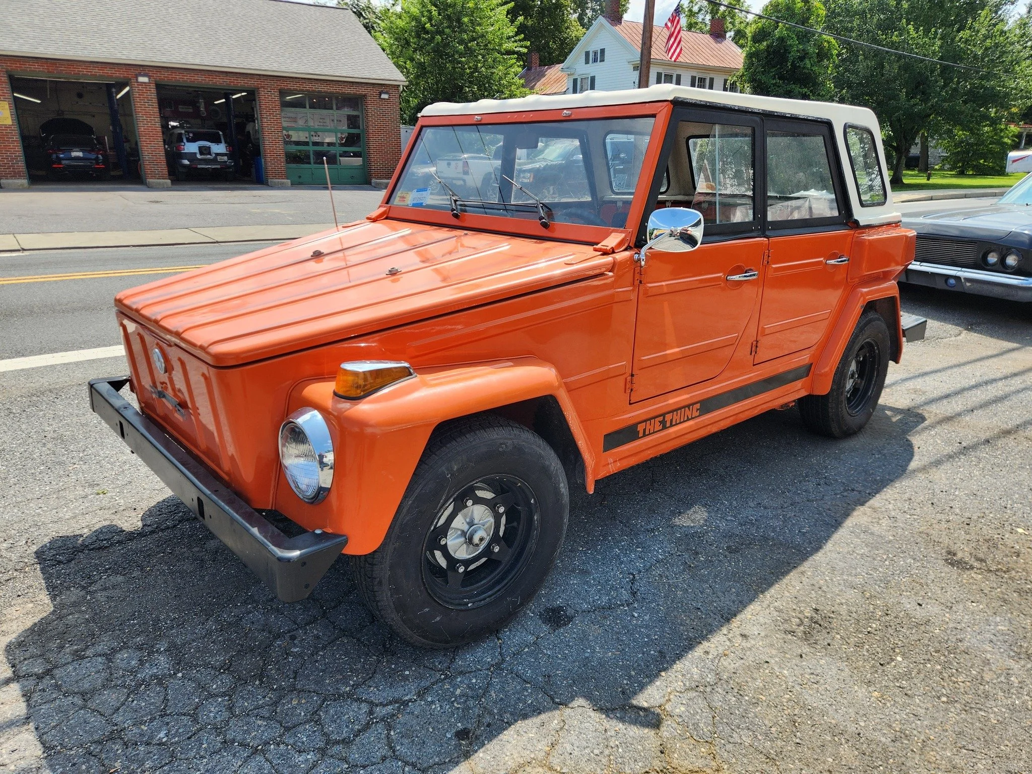 A vintage orange and white off-road vehicle parked on a street, with a black stripe on the lower side that reads "THE THING." In the background, a brick building with an open garage and a few other cars are visible.