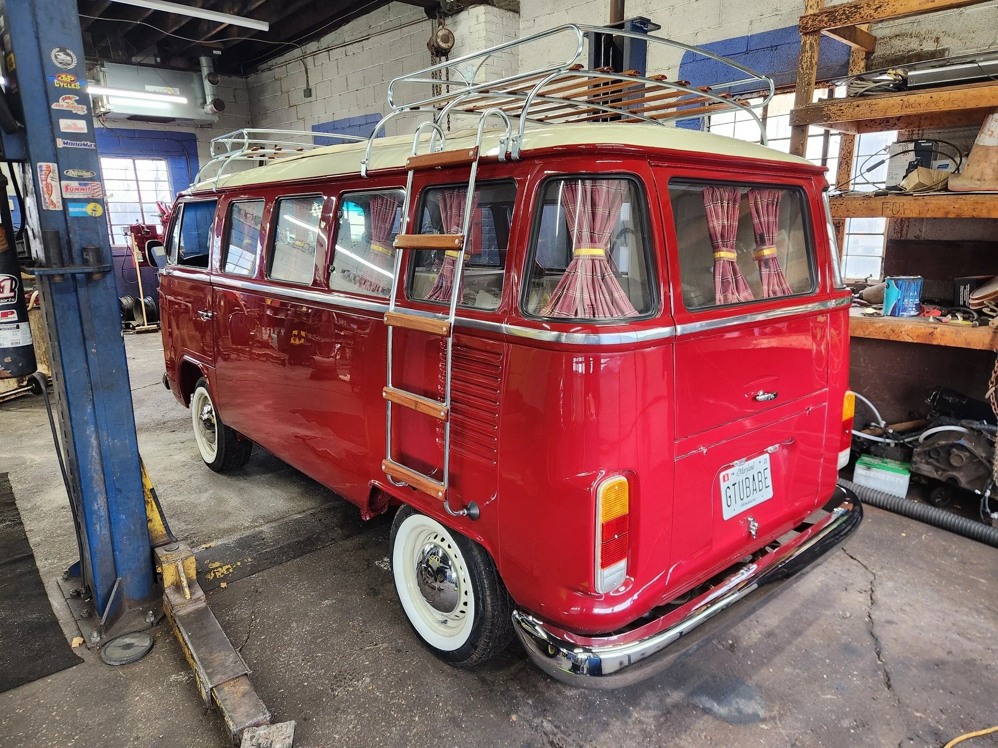 A red vintage van with a white roof, featuring a ladder on the side, is parked inside a workshop. The van has small curtains on the windows and a custom license plate that reads 'GTUBABE.' The workshop has various tools and parts on shelves and a blu