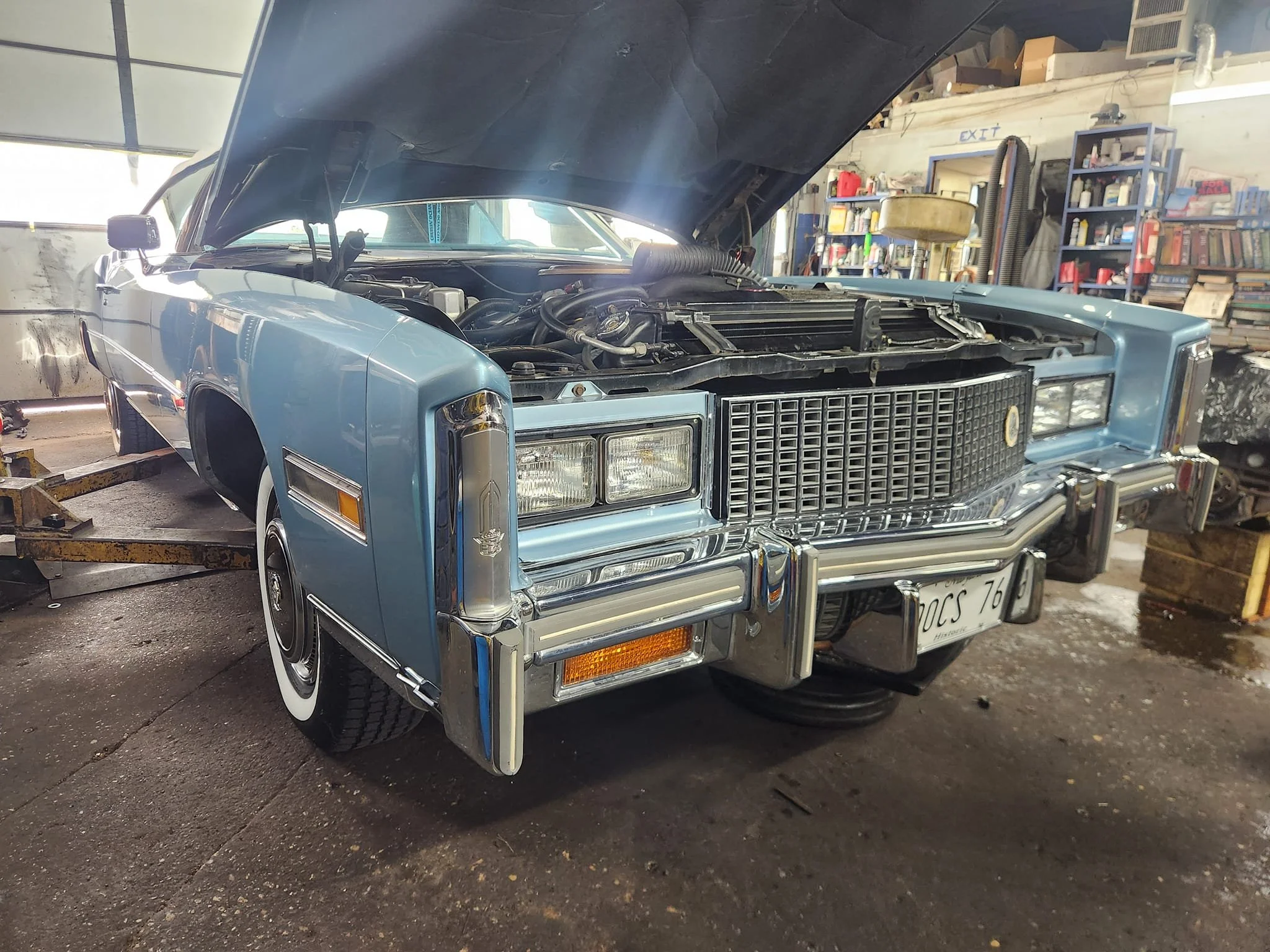 A vintage light blue Cadillac with its hood open in a garage or workshop, with tools and shelves in the background.