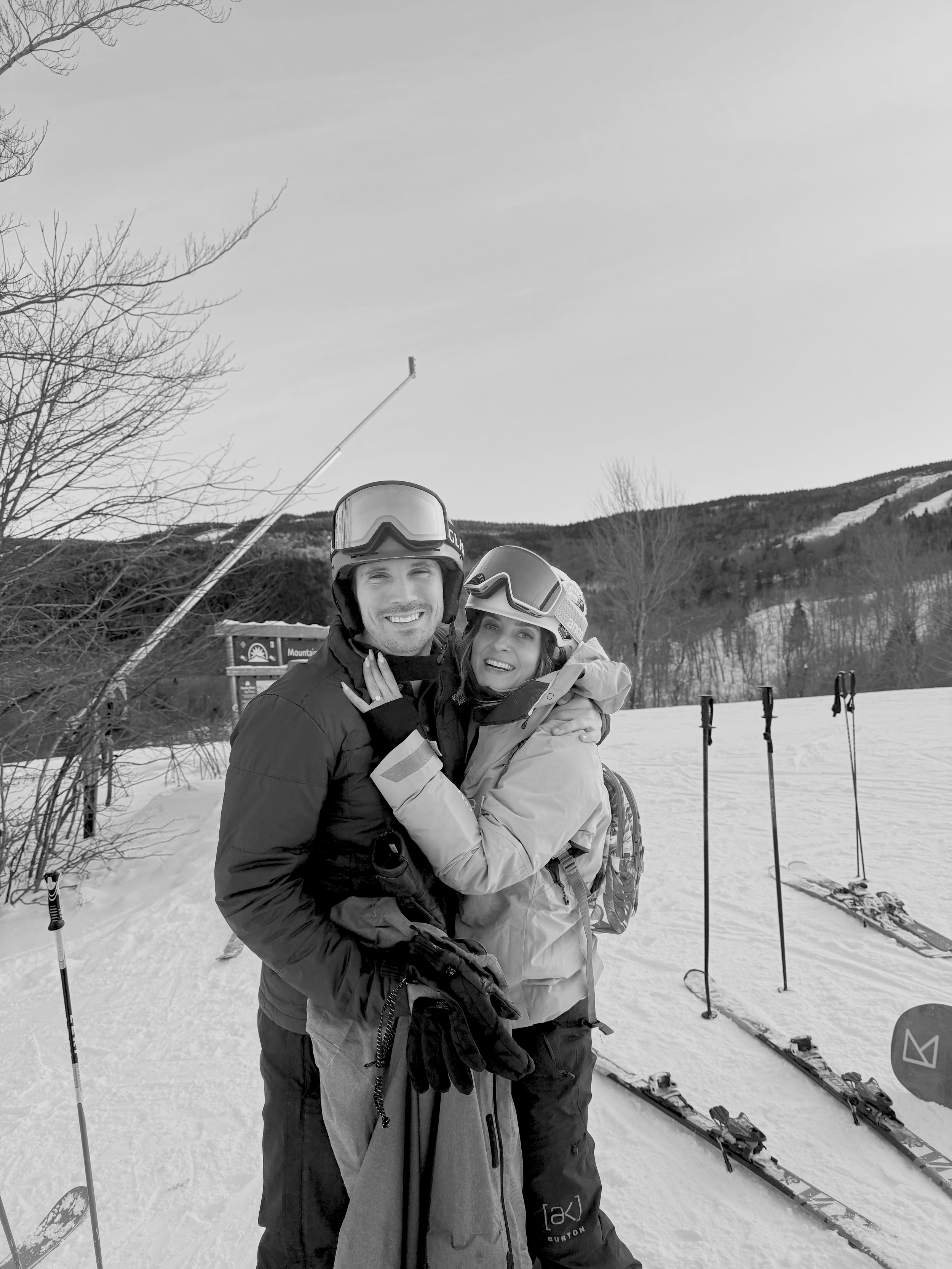 A couple dressed in ski gear embracing on a snowy mountain with skiing equipment behind them.