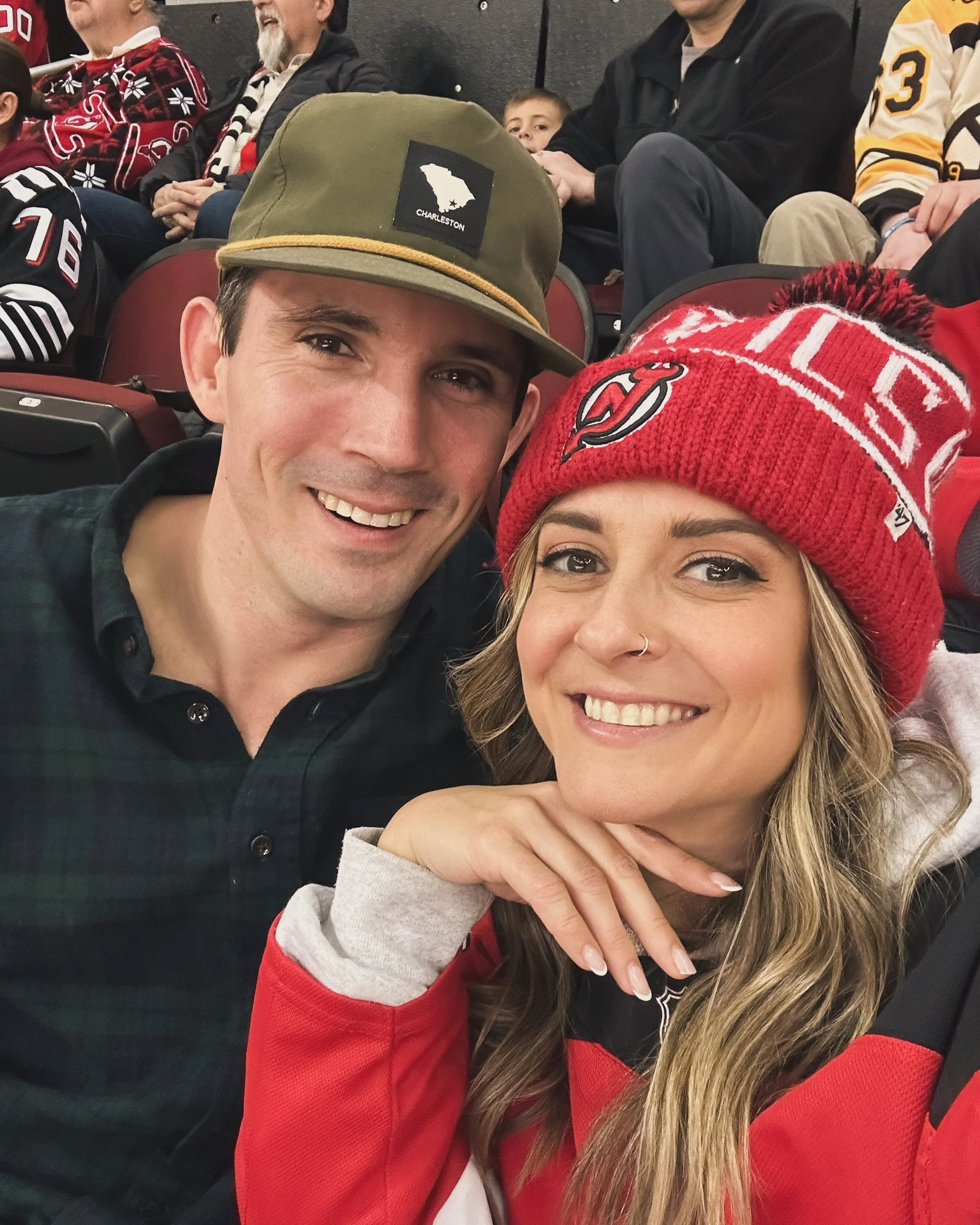 A smiling man and woman taking a selfie at a sports venue, with the man wearing a green baseball cap featuring a map of South Carolina and the woman wearing a red knit hat with a sports team logo, surrounded by other spectators.