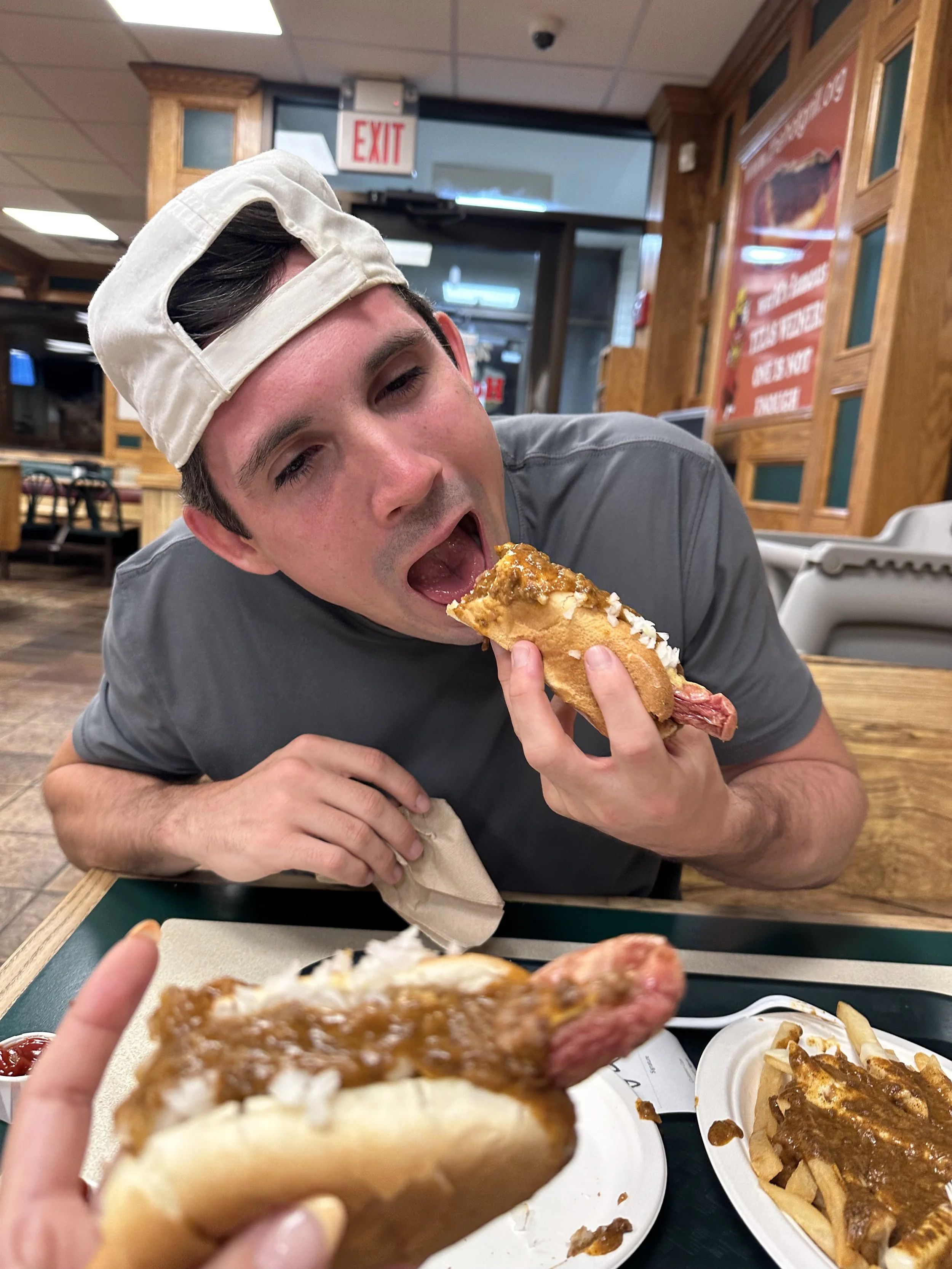 A man wearing a baseball cap is eating a hot dog in a casual restaurant, with another hot dog and fries on the table.