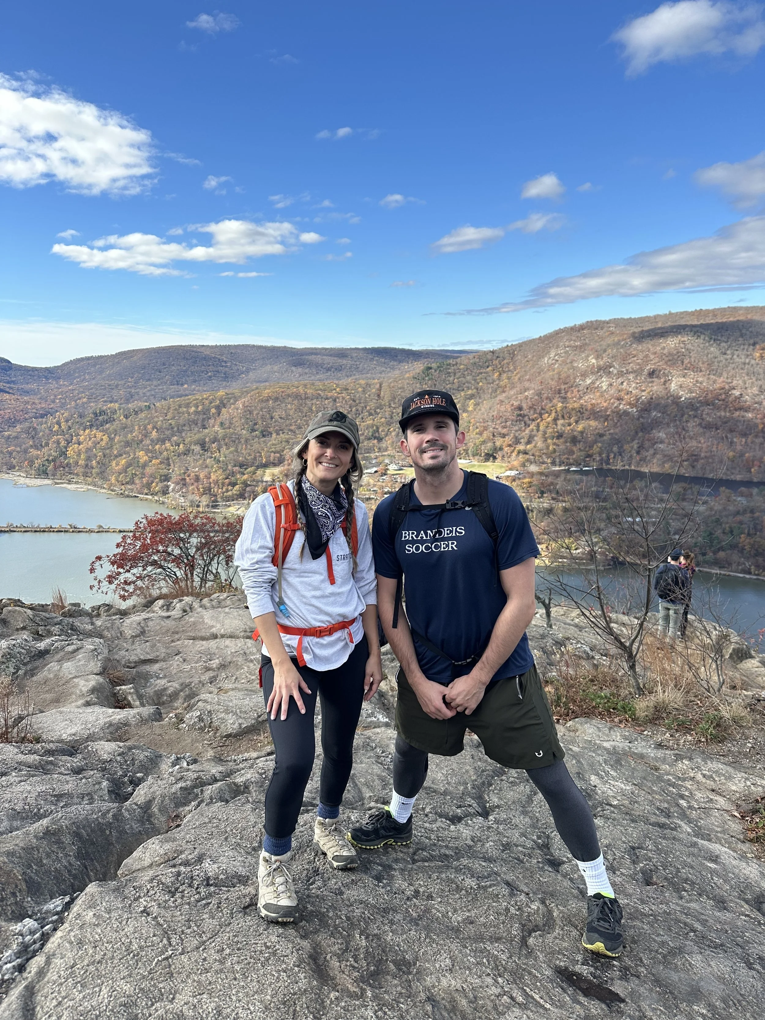 Two hikers, a woman and a man, standing on a rocky hilltop with a scenic view of a river and mountains in the background on a clear day.