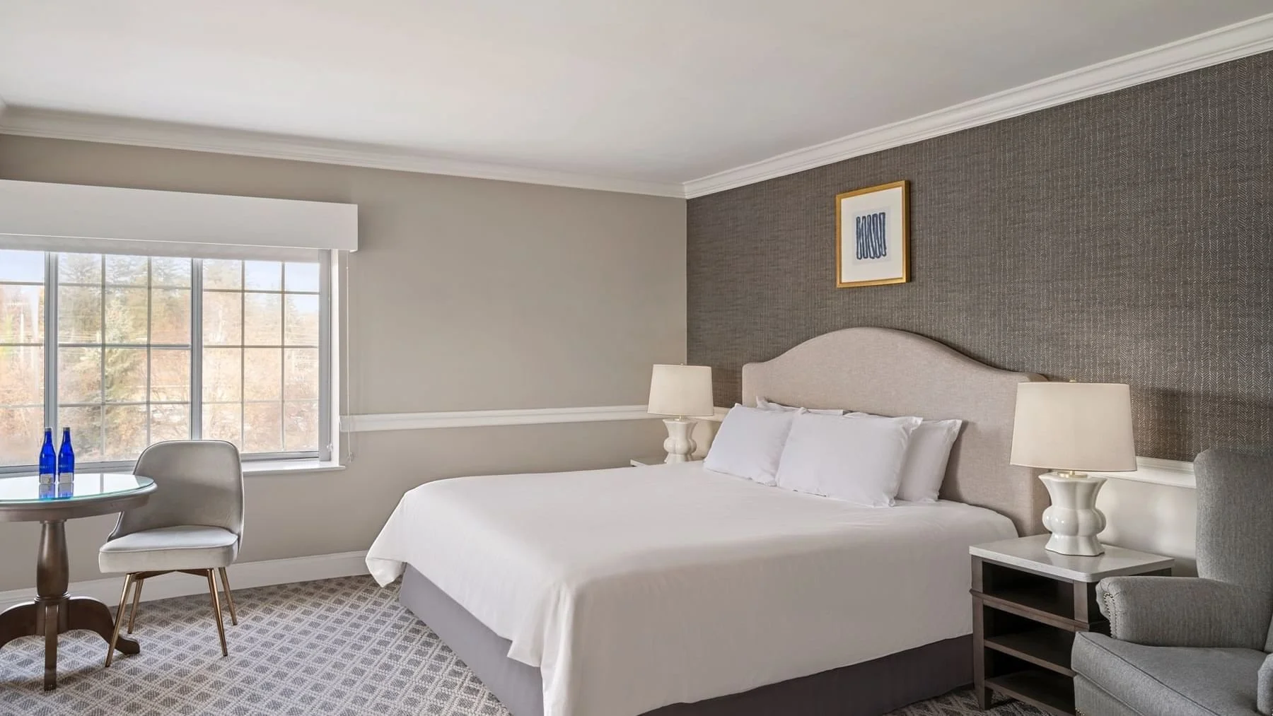 Hotel bedroom with a large bed, nightstand lamps, window with a view, and sitting area with a small table and two chairs.