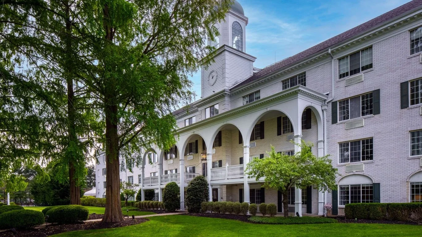 A white multi-story building with a clock tower, surrounded by green trees and manicured bushes, under a partly cloudy sky.