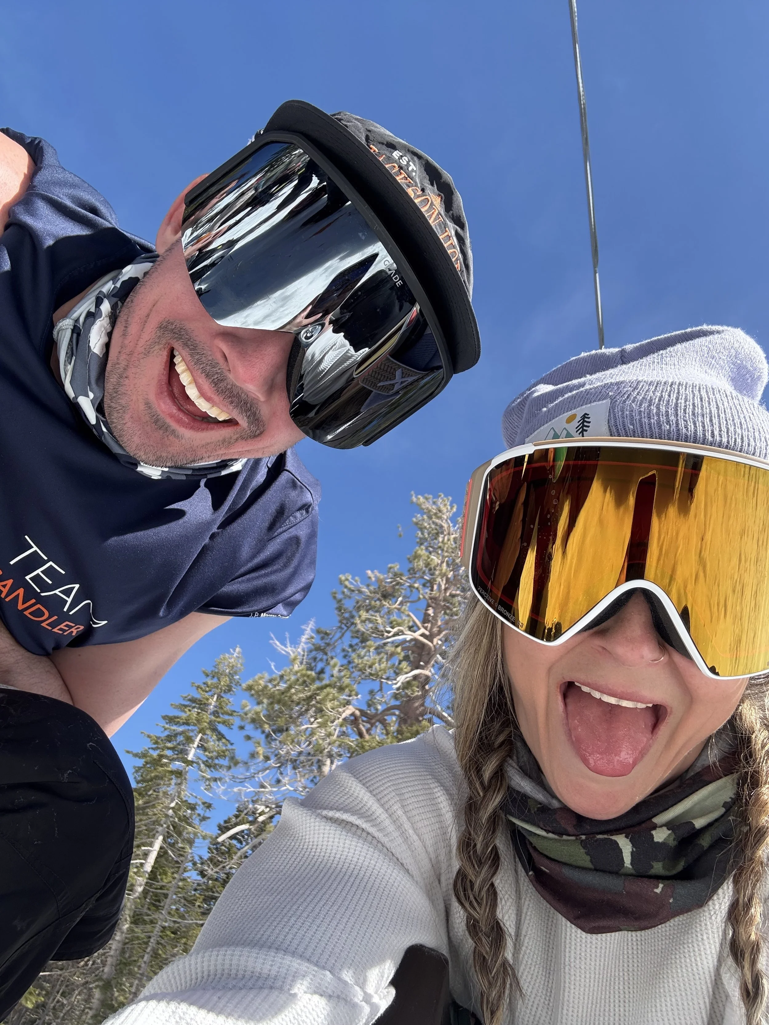 Two people in snow gear and ski goggles taking a selfie outdoors on a sunny ski day with snow-covered trees and a blue sky in the background.