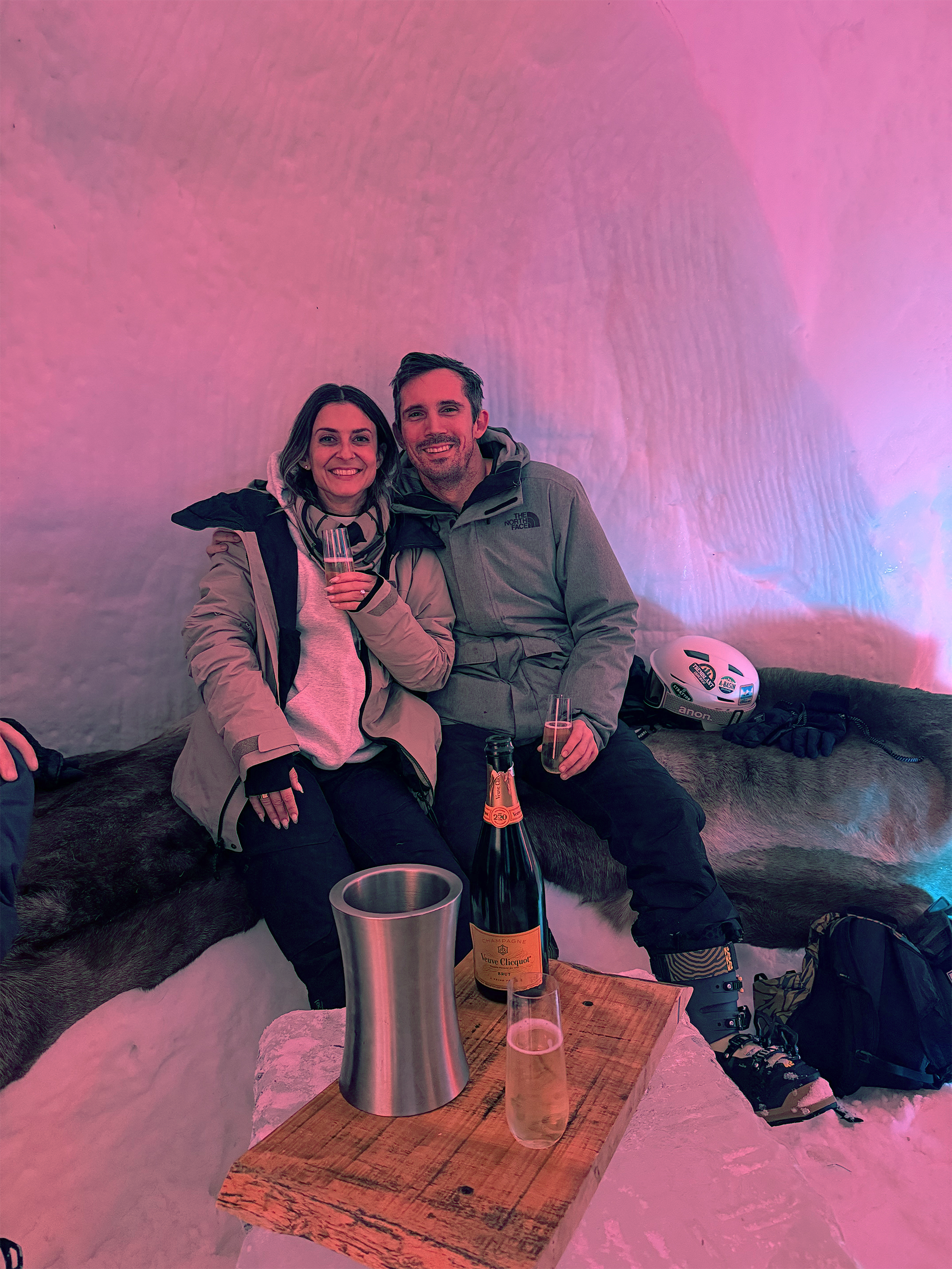 A smiling woman and man sitting on a hide covered with animal fur, holding champagne glasses. The woman is wearing a beige jacket and gloves, and the man is in a gray jacket and skiing boots. There is a bottle of champagne and a glass on a small wooden table in front of them, a snow helmet and gloves on the fur, and a backpack on the snow. The background has pink and purple lighting.
