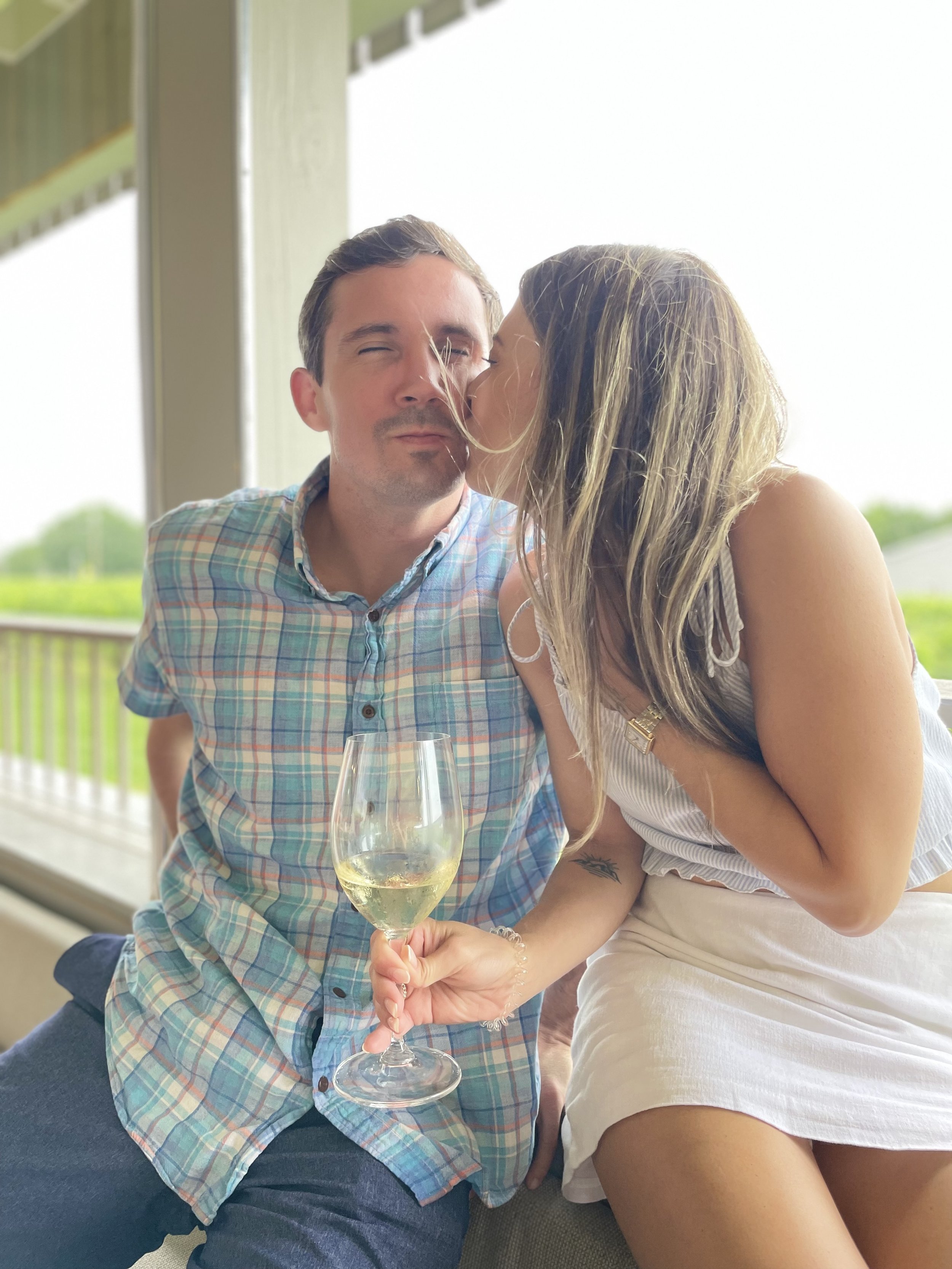 A woman gives a kiss on the cheek to a man holding a glass of white wine, on a balcony with a green landscape in the background.