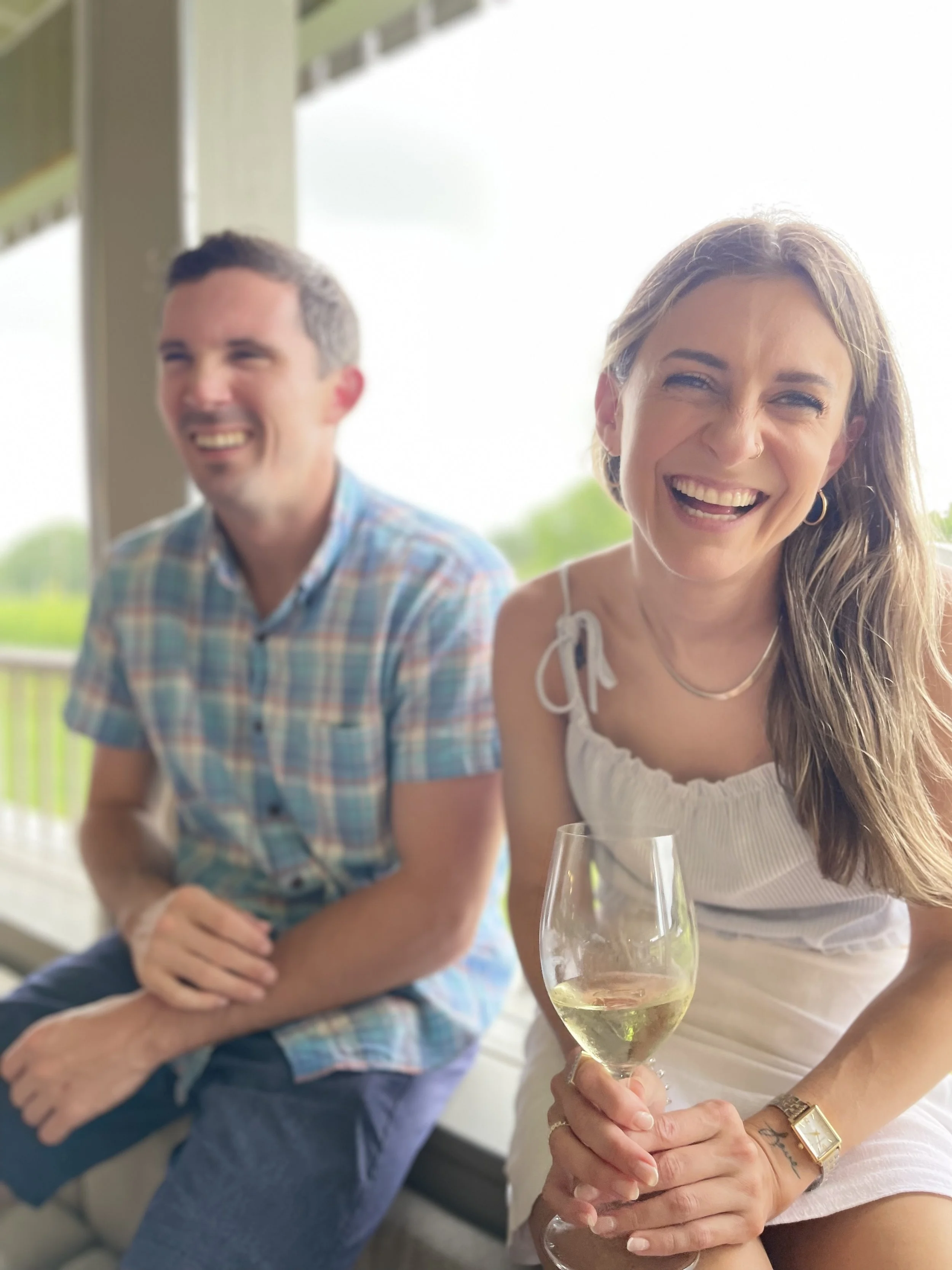 A woman with long hair holding a glass of white wine, smiling, sitting next to a man in a plaid shirt on a porch with a green landscape in the background.