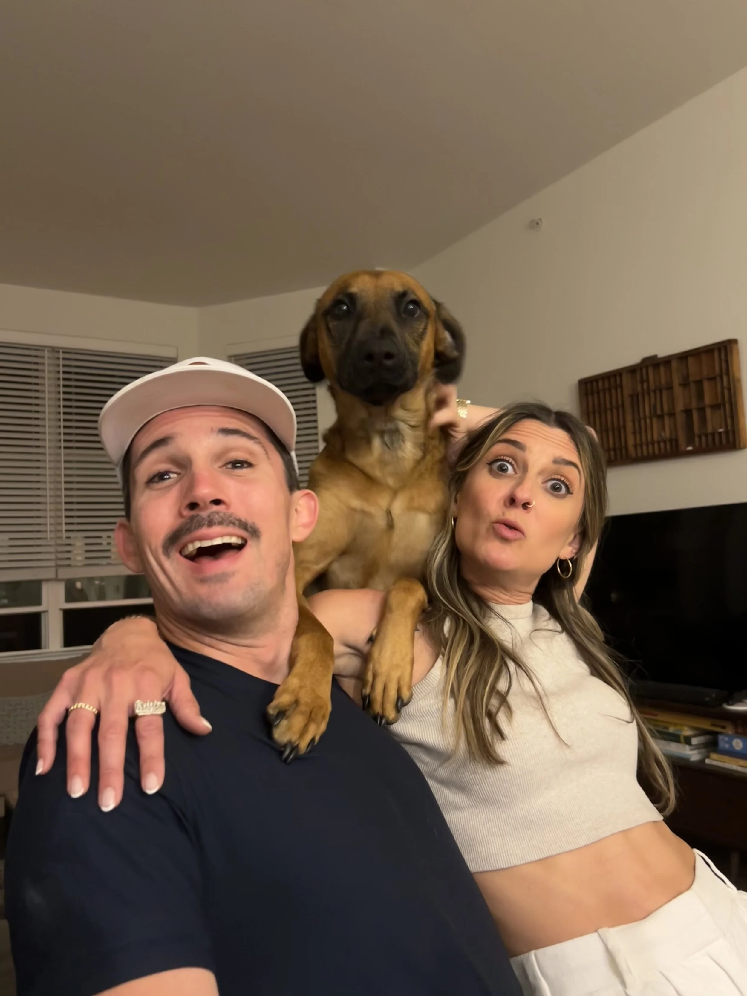 A man, woman, and a dog posing for a photo indoors. The man is wearing a white cap and a black shirt, smiling; the woman has long wavy hair, is wearing a crop top, and making a playful face. The dog, a medium-sized brown with black face, is held up between them, looking at the camera.