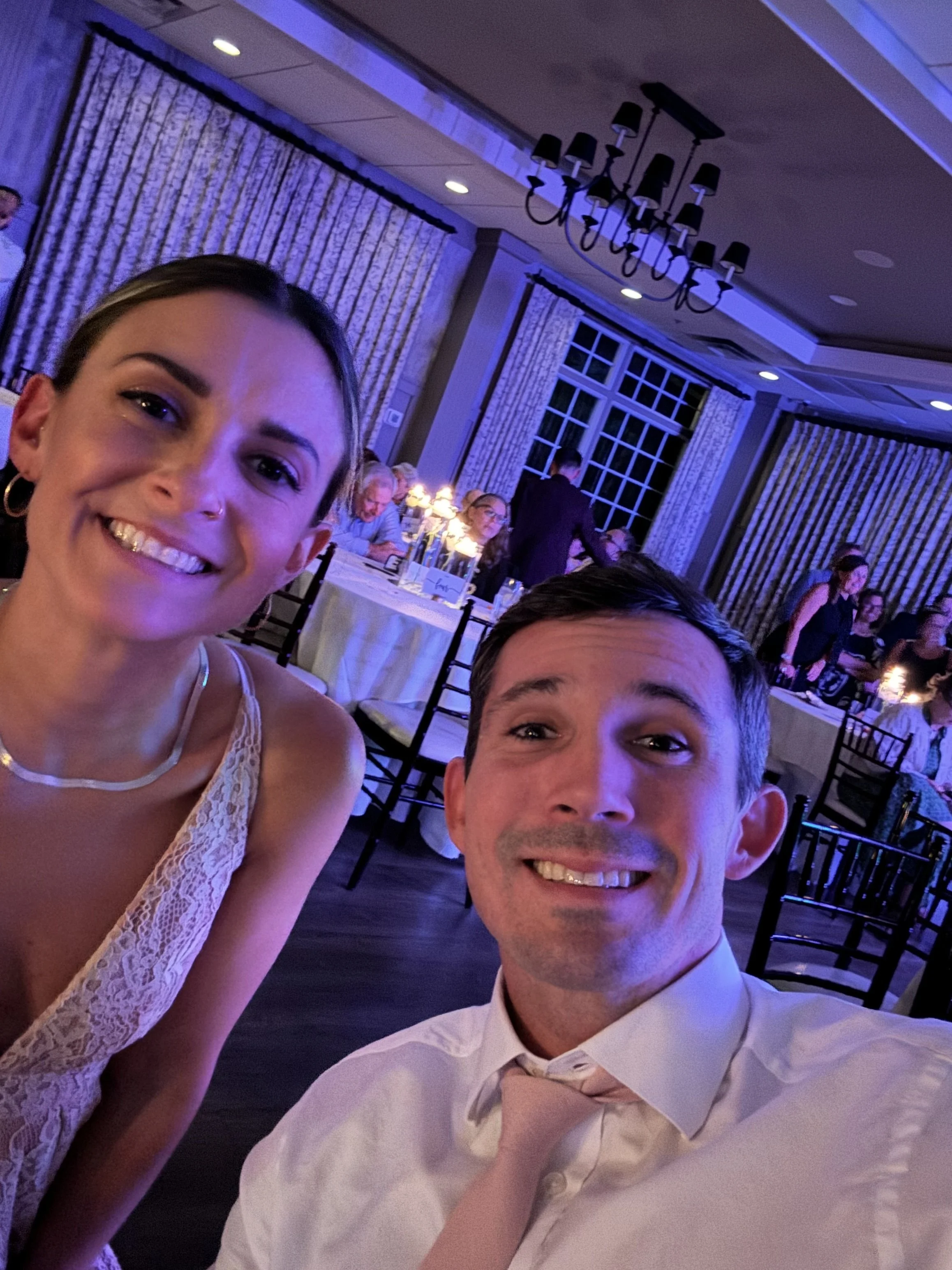 A smiling woman and man taking a selfie at a formal event or celebration, with tables and guests in the background, and purple lighting in the room.