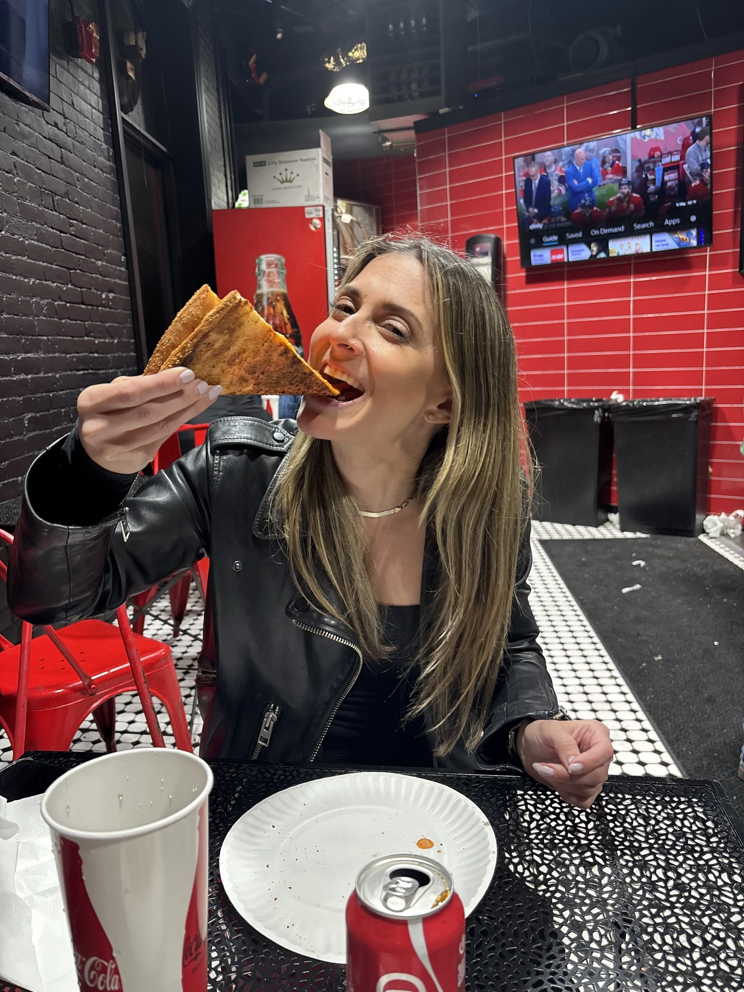 A woman with long light brown hair, wearing a black leather jacket, is smiling and about to take a bite of a slice of pizza in a restaurant with a black wall and red tiled wall background. On the table are two Coca-Cola cans, an empty paper cup, and a white paper plate with some food remnants.