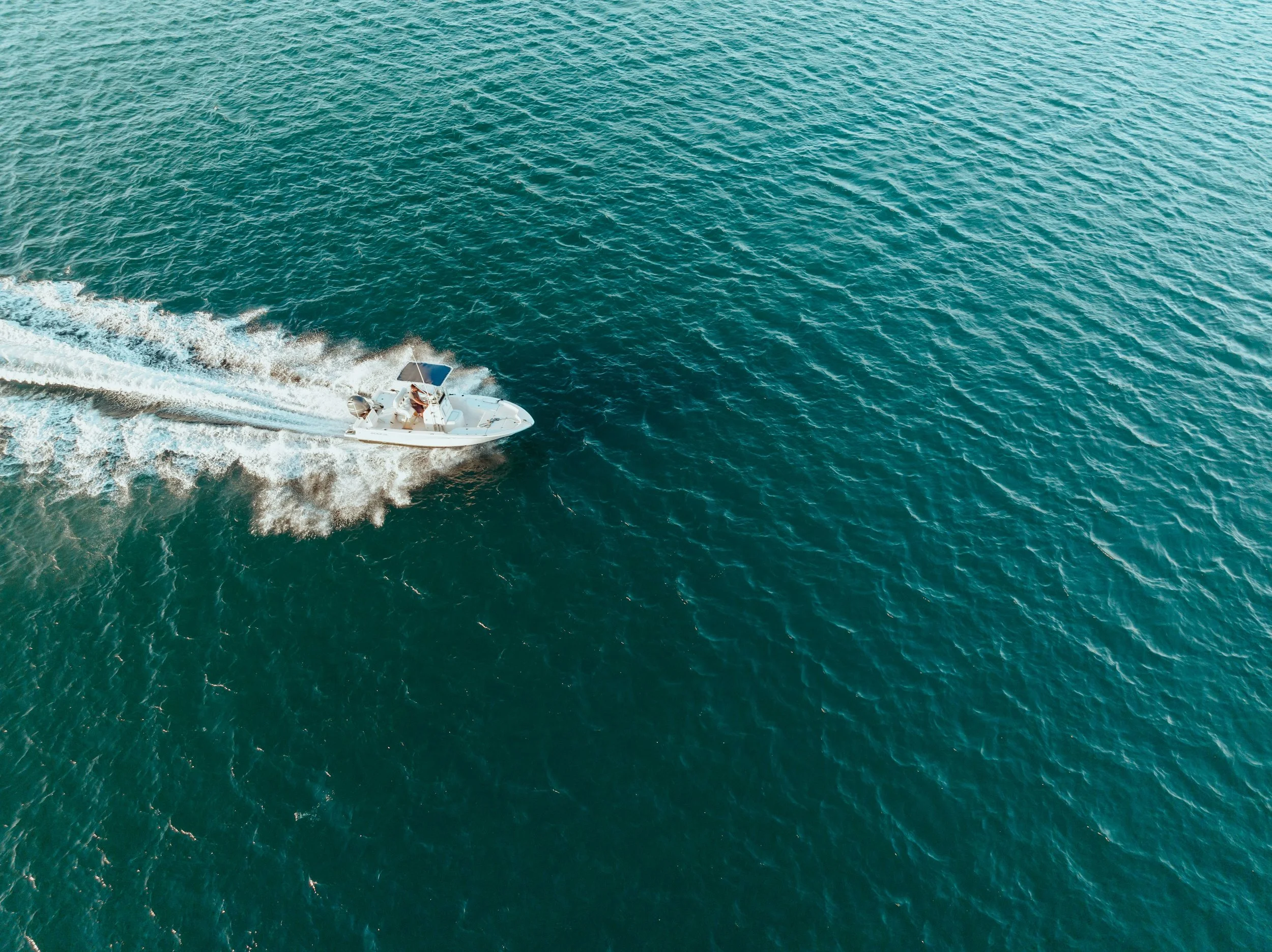 An aerial view of a small white boat with two people on board speeding across the water, creating a wake behind it.