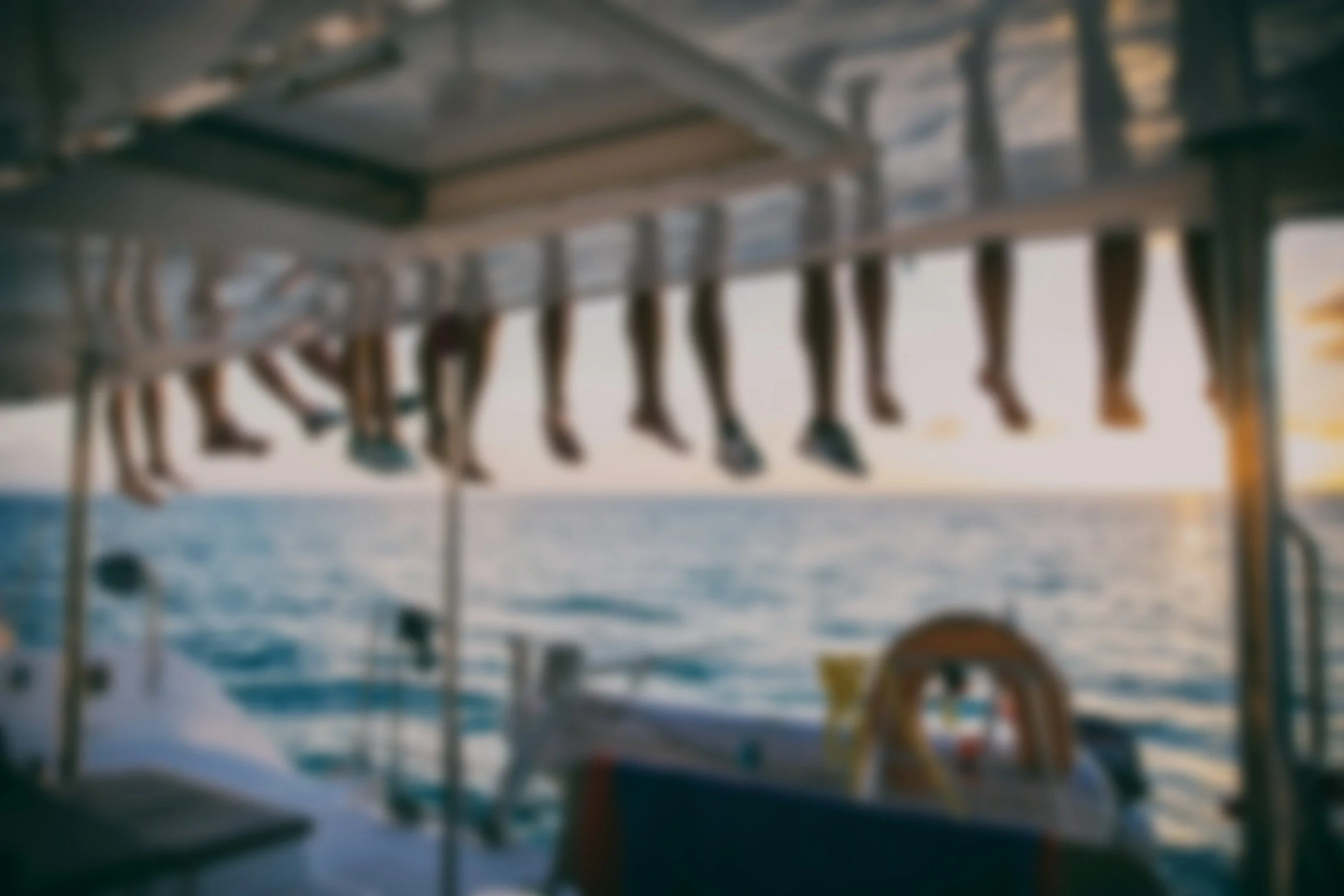 Blurred photo of a boat viewed from the inside, showing human legs hanging off the side of the boat, with the ocean and sunset in the background.