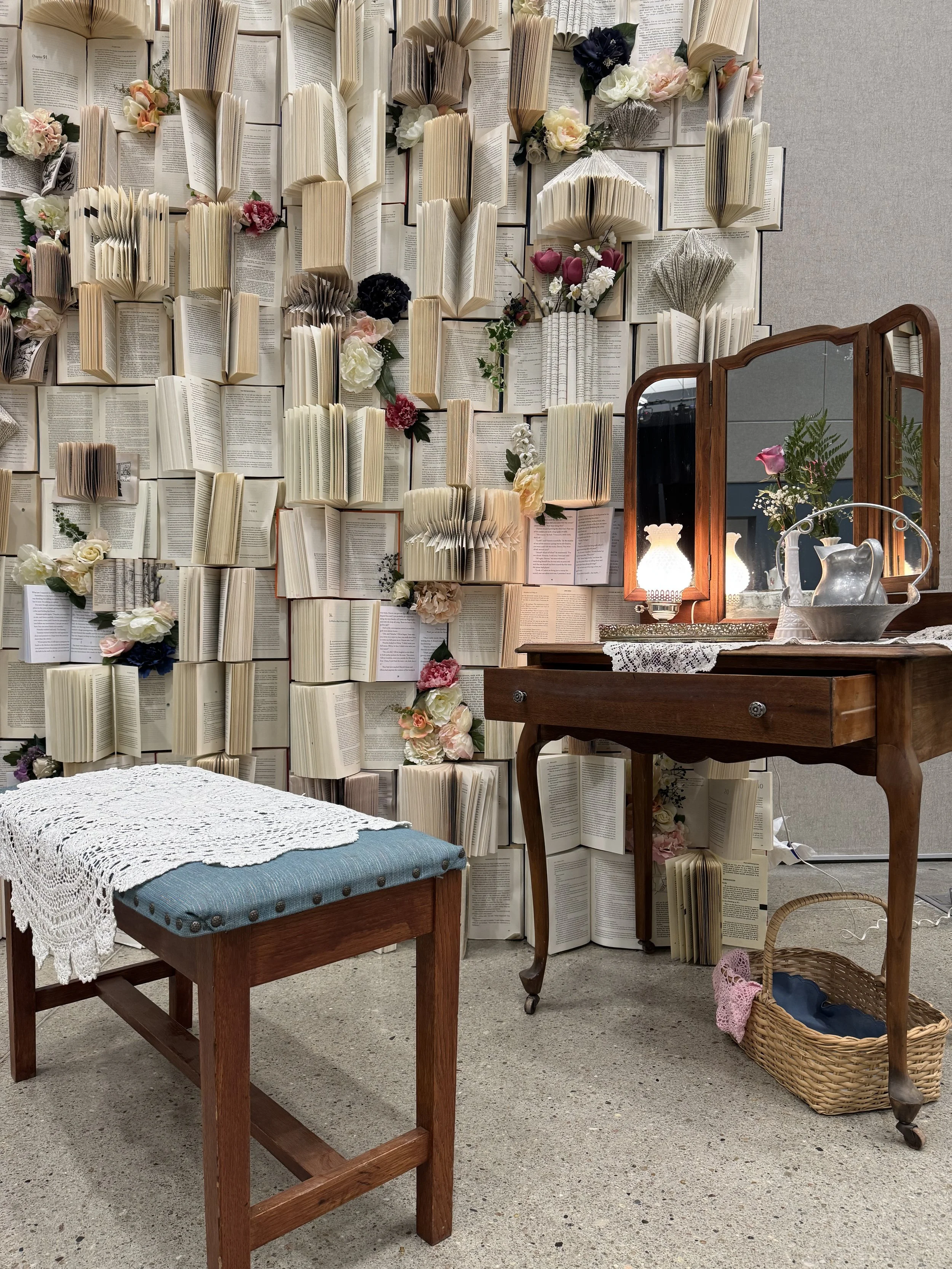 An interior setting with a wall decorated with numerous open books and flowers, a vintage wooden dressing table with a mirror, a small table with a crocheted lace cloth, and a wicker basket on the floor.