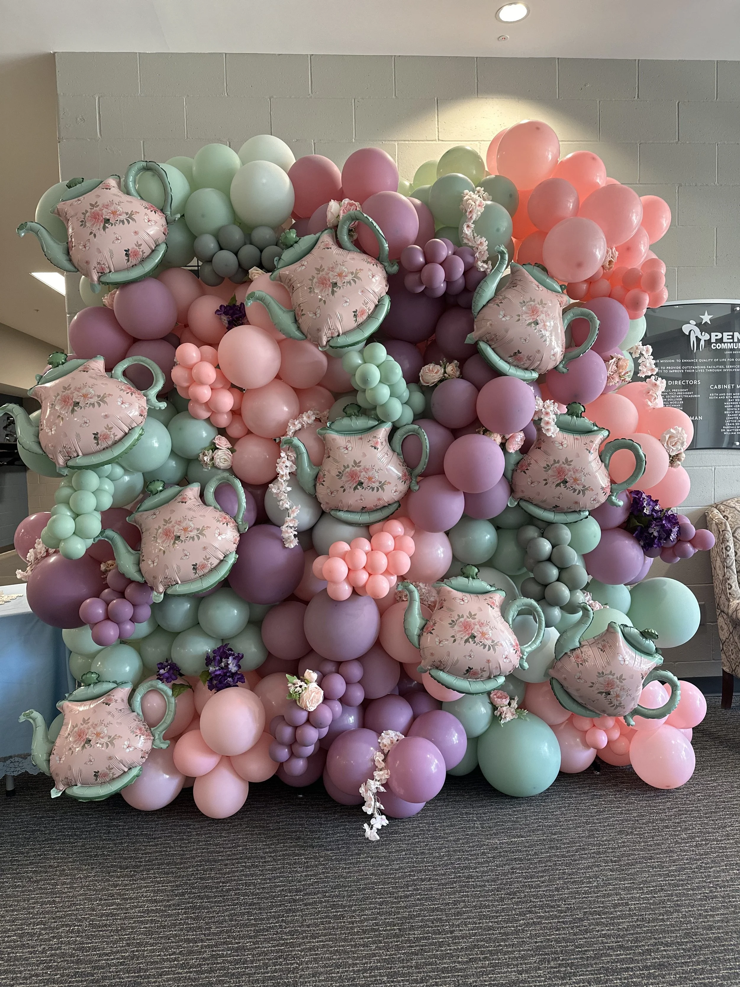Decorative balloon arrangement with pastel-colored balloons, teapot-shaped balloons, and small flowers, creating a garden party theme.