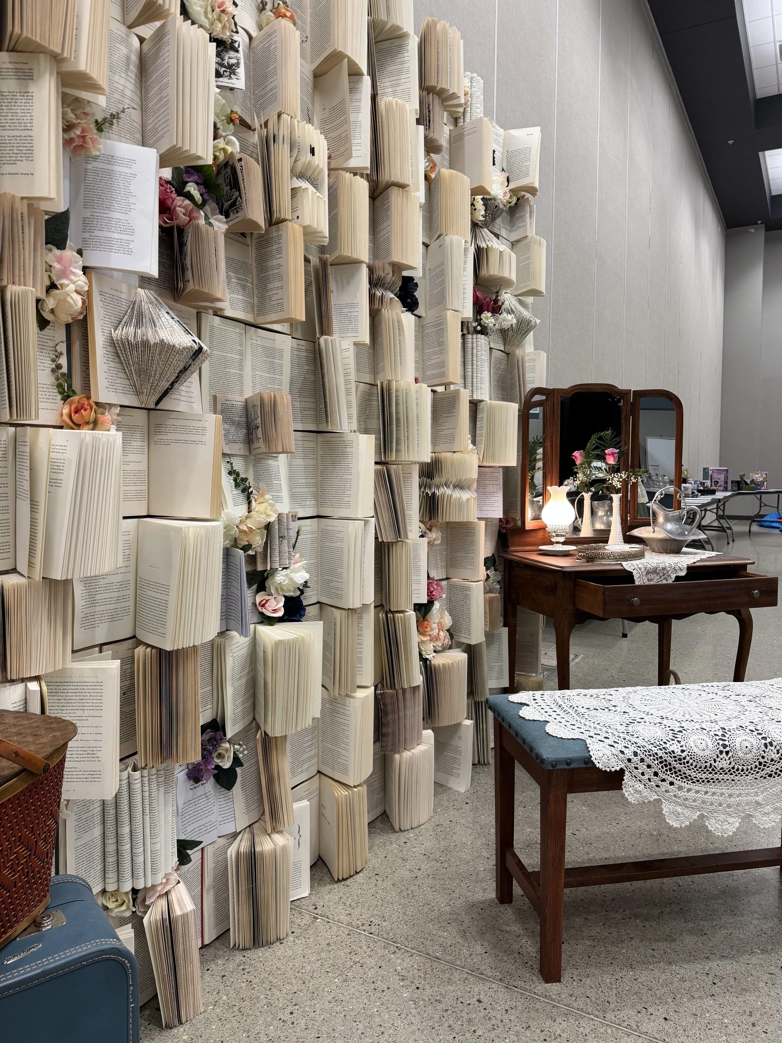 Wall made of open books with flowers and a mirror, vintage wooden vanity table with lamps and flowers, and a table with a lace tablecloth.