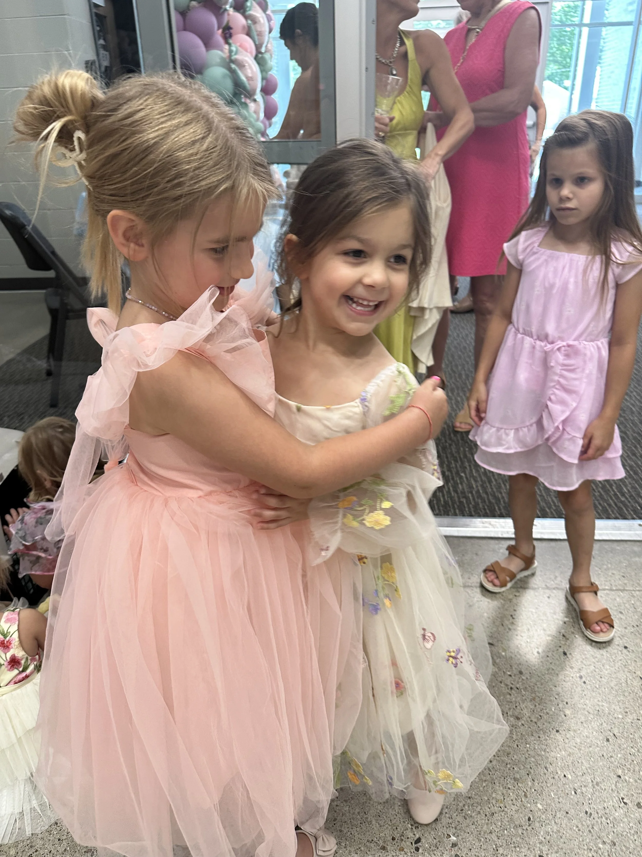 Three young girls in pastel dresses at a party, with two women in the background near balloons. The girls are smiling and interacting, with one girl in a peach dress hugging another girl in a floral-patterned dress.