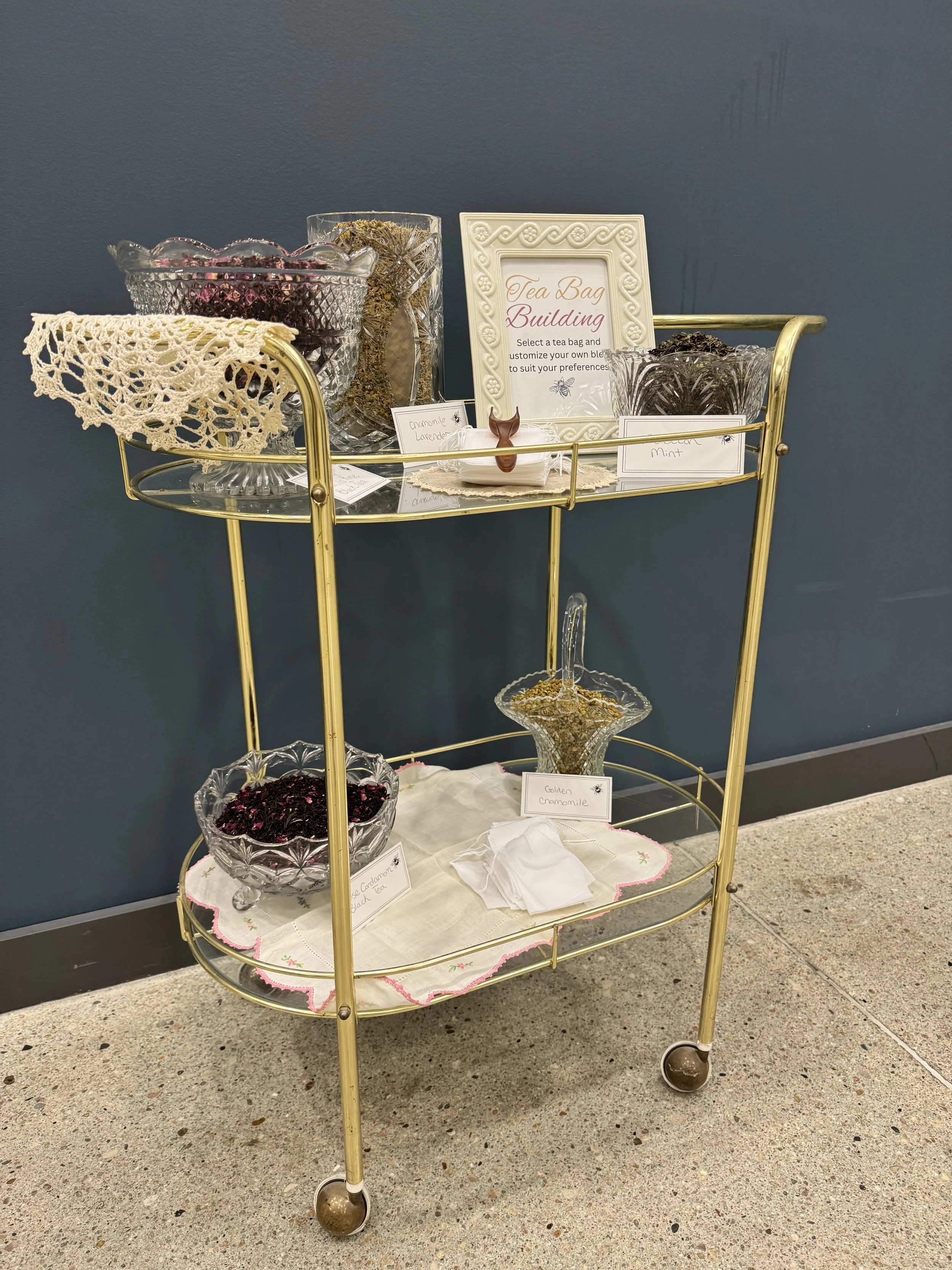A gold-colored beverage cart with glass bowls of dried herbs, flowers, and tea ingredients, and a framed sign about tea bag building, set against a dark blue wall.