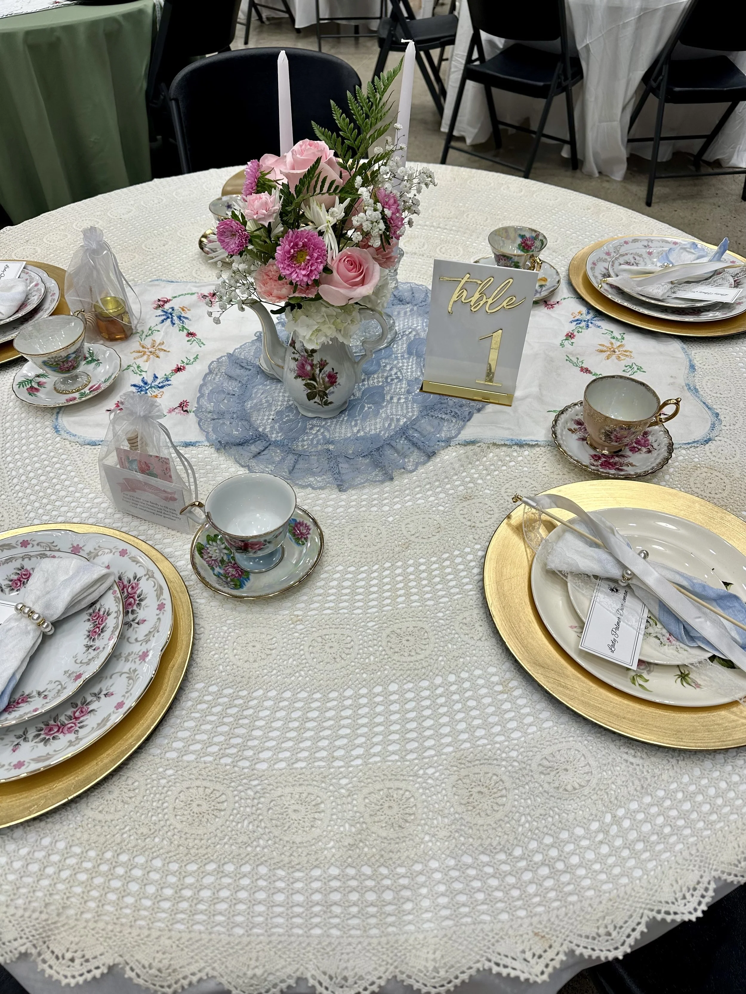 A round dining table set for a tea party or brunch with floral-patterned china, gold-rimmed plates, and teacups. The centerpiece is a pink flower arrangement with candles, and there is a table number card indicating 'Table 1'.