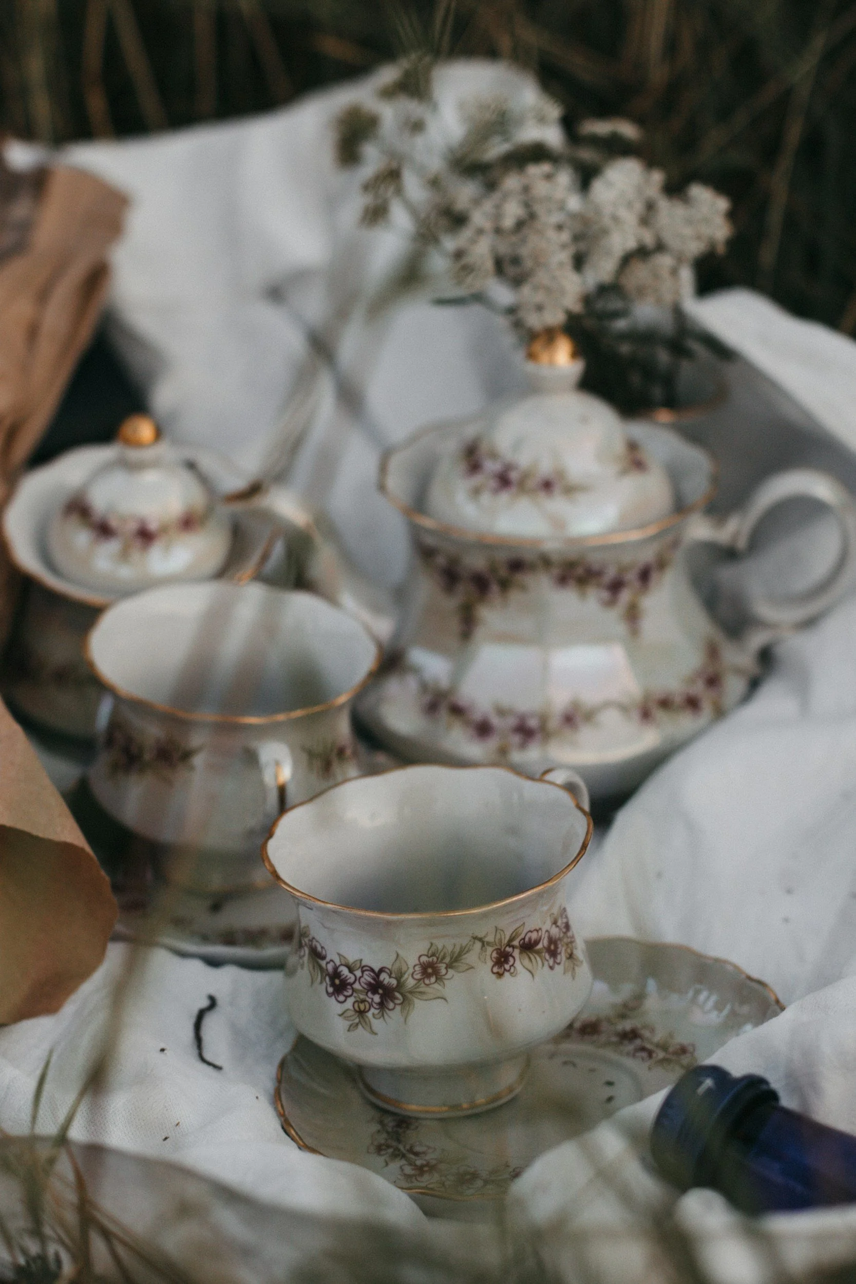 Vintage porcelain tea set with floral patterns and gold trim, placed on a white fabric with dried flowers in the background.