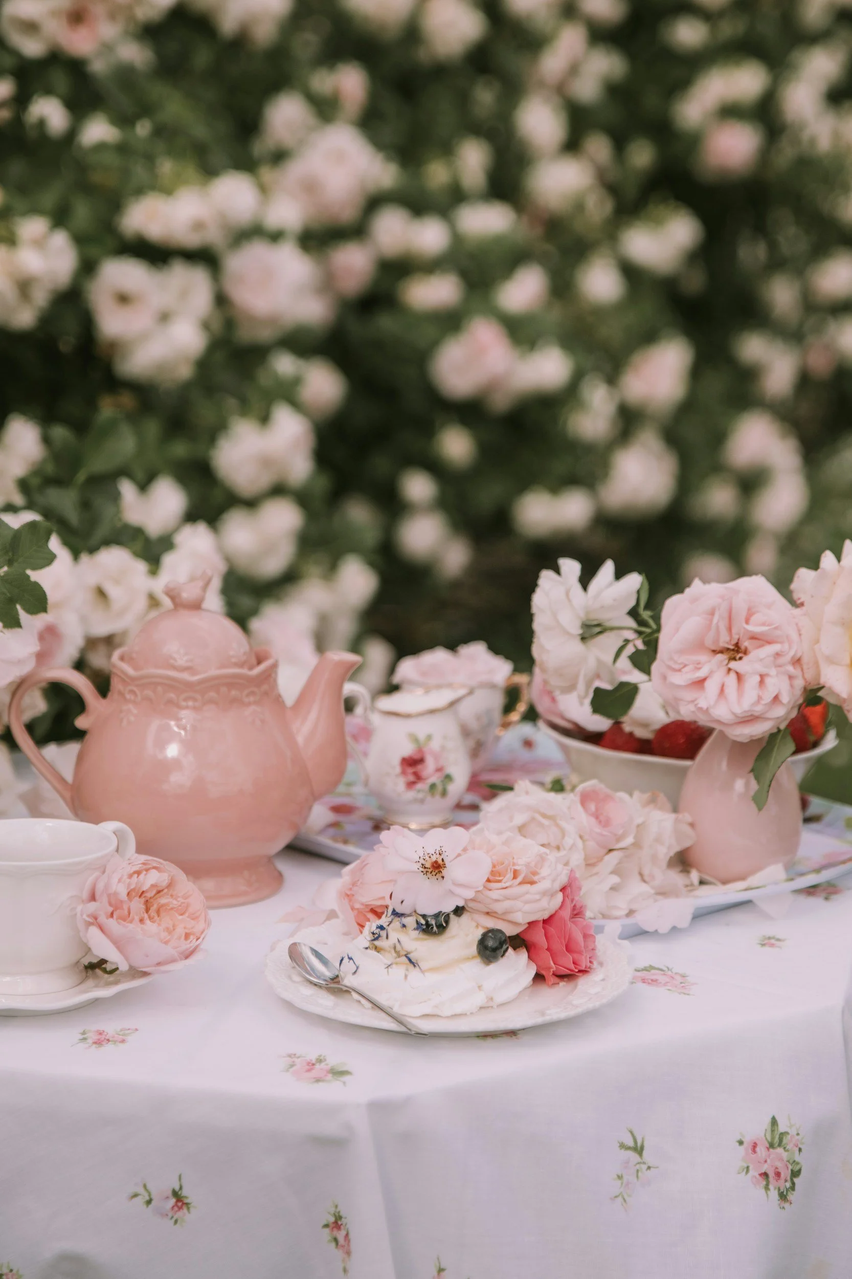 Tea set with pink teapot, cups, and plates on a floral tablecloth, surrounded by pink and white flowers and greenery.