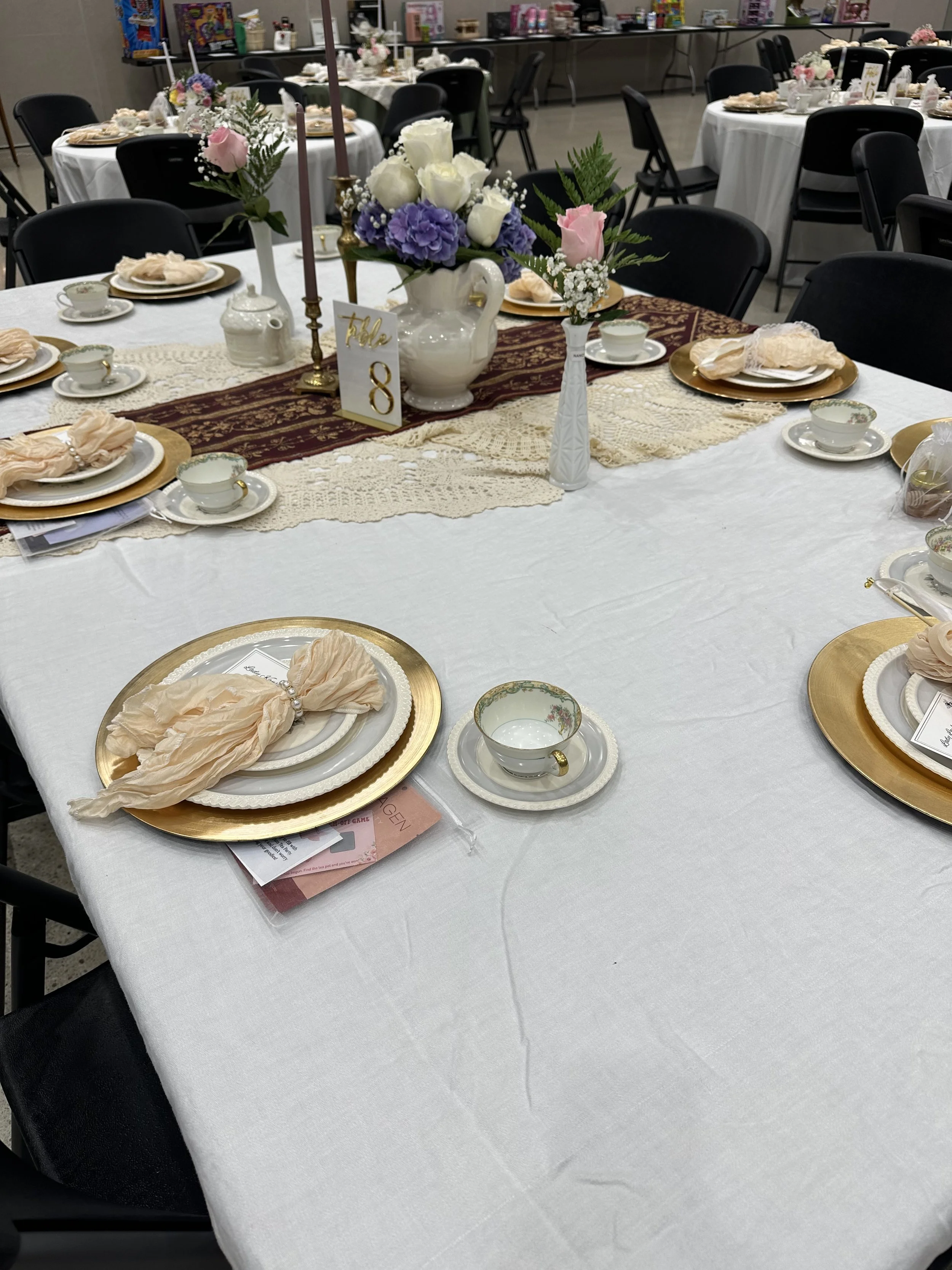A decorated banquet table set for a special event, featuring elegant china teacups and saucers, gold chargers, cream-colored cloth napkins tied with pearl pins, a floral centerpiece with white, purple, and pink flowers, and a table number sign marked