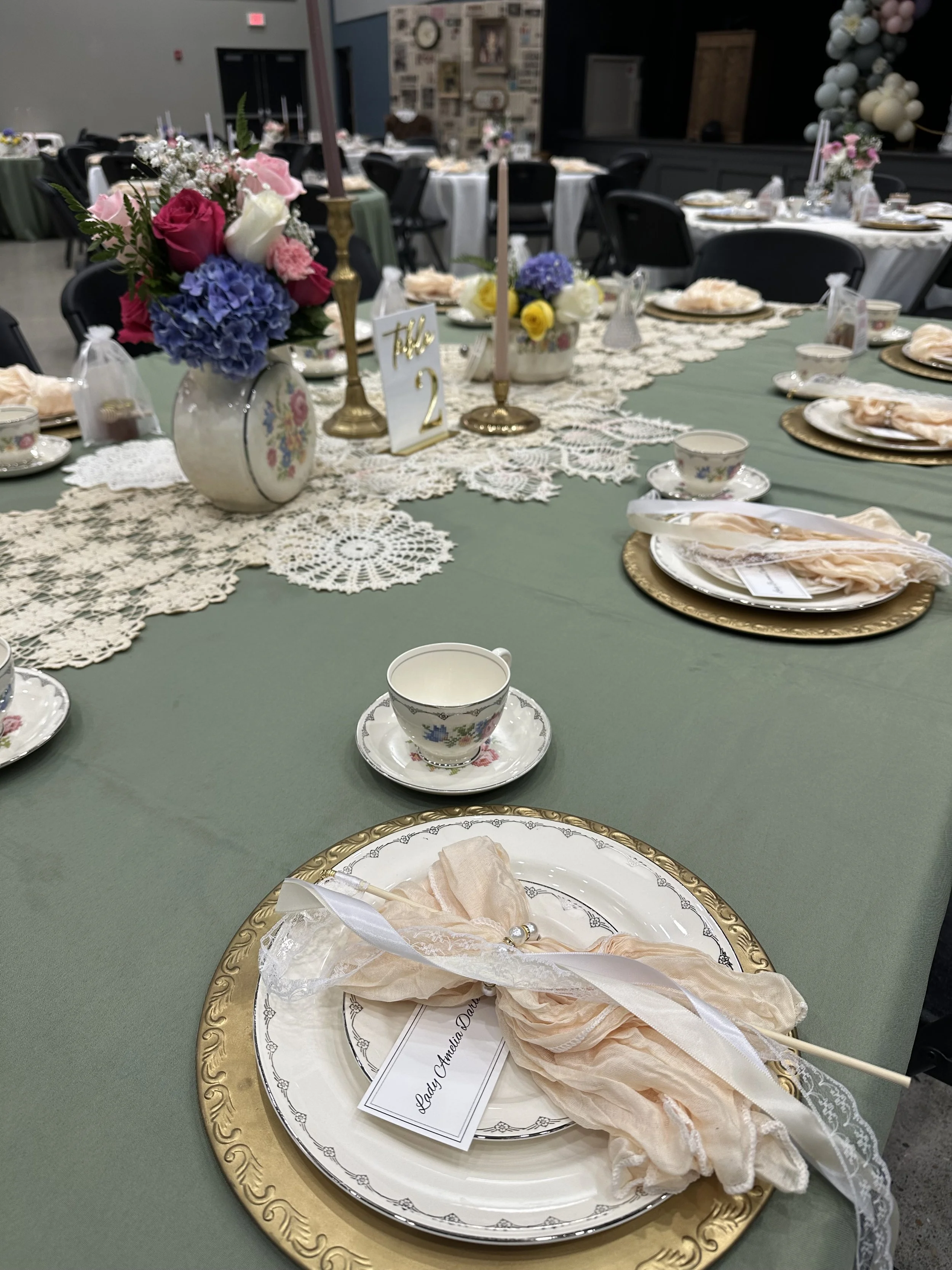 Elegant table setting for a celebration with floral arrangements, lace table runners, teacups, saucers, gold chargers, and place cards.